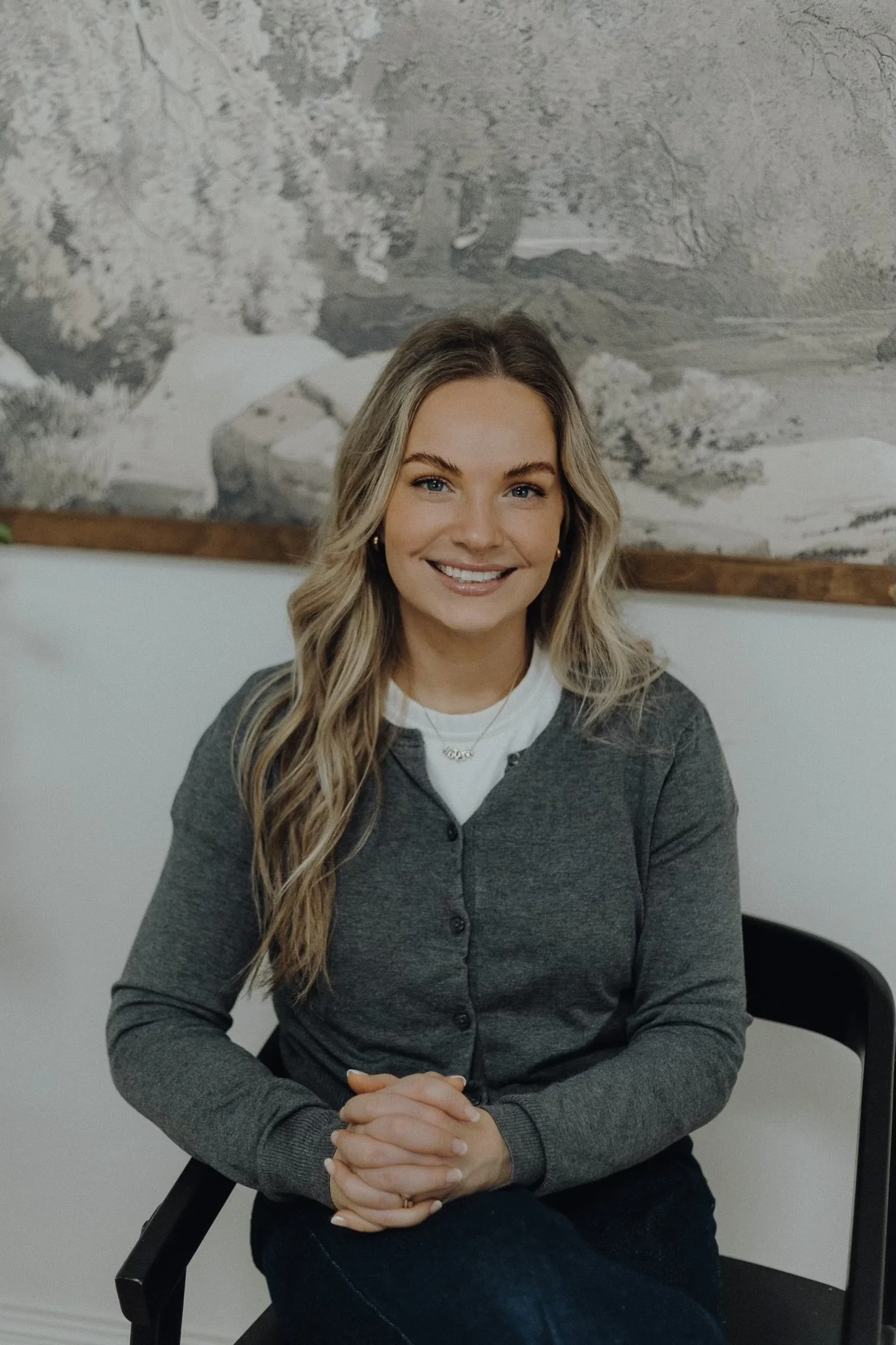 A woman with long, wavy blonde hair, wearing a gray cardigan and a white shirt, is smiling while sitting on a black chair with her hands clasped in her lap, in front of a wall with a gray landscape artwork.