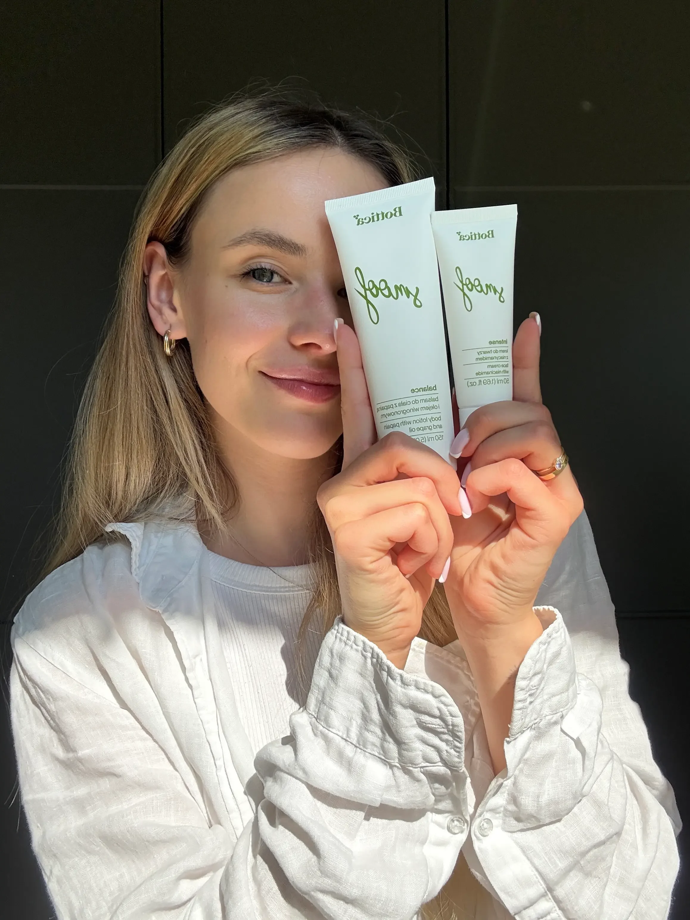 A young woman with blonde hair and a white shirt holding two tubes of skincare product labeled 'berry' and posing with a slight smile against a black background.