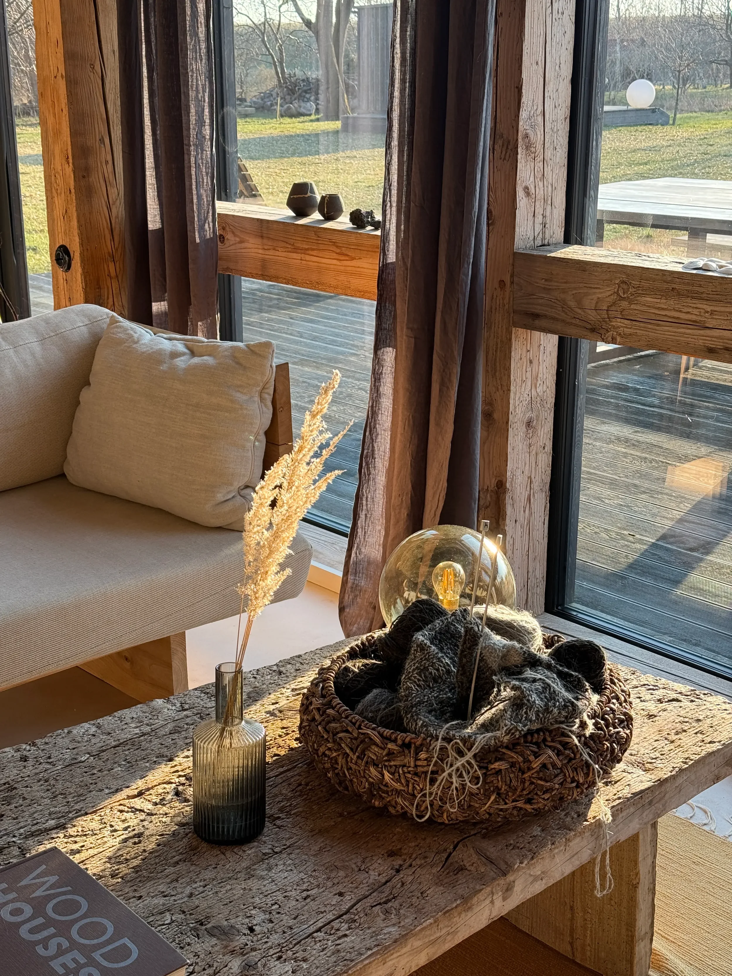 Sunlit rustic living room with a beige sofa and a weathered wooden table holding a dark glass vase with dried pampas grass, a woven basket filled with yarn and knitting needles, and a spherical decorative light. Large glass windows with brown curtains reveal a grassy outdoor area.