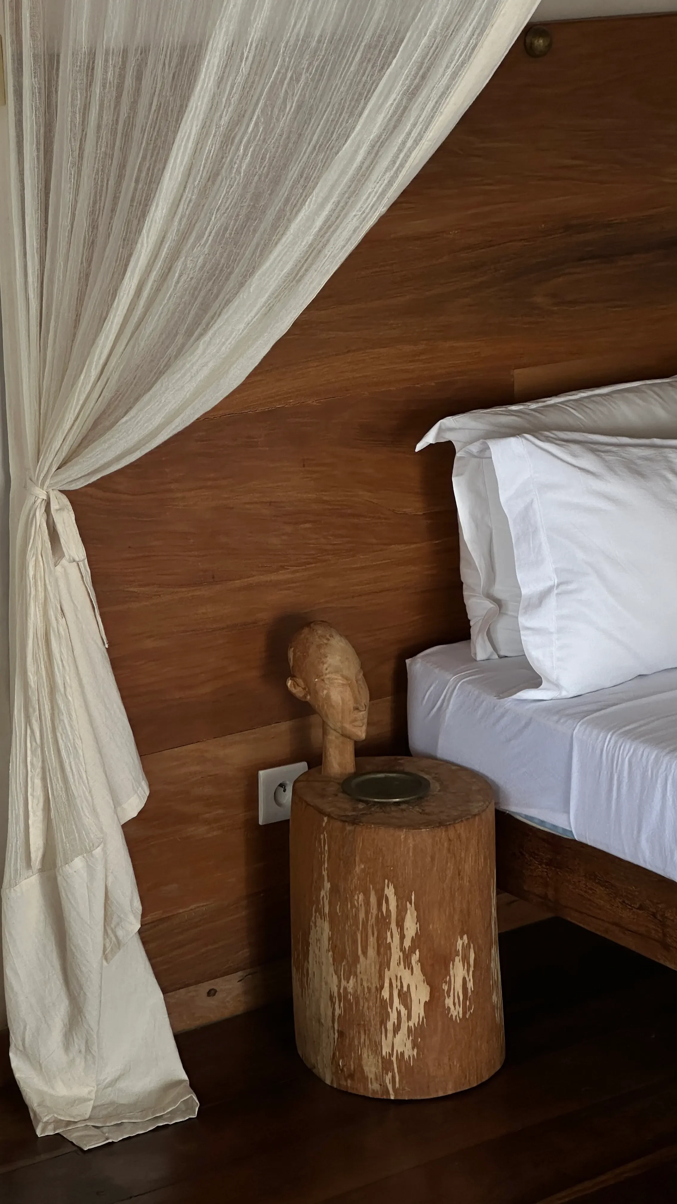 A bedroom corner with a wooden headboard, white bed sheets, a bedside table made from a tree trunk, and a carved wooden bust sculpture.