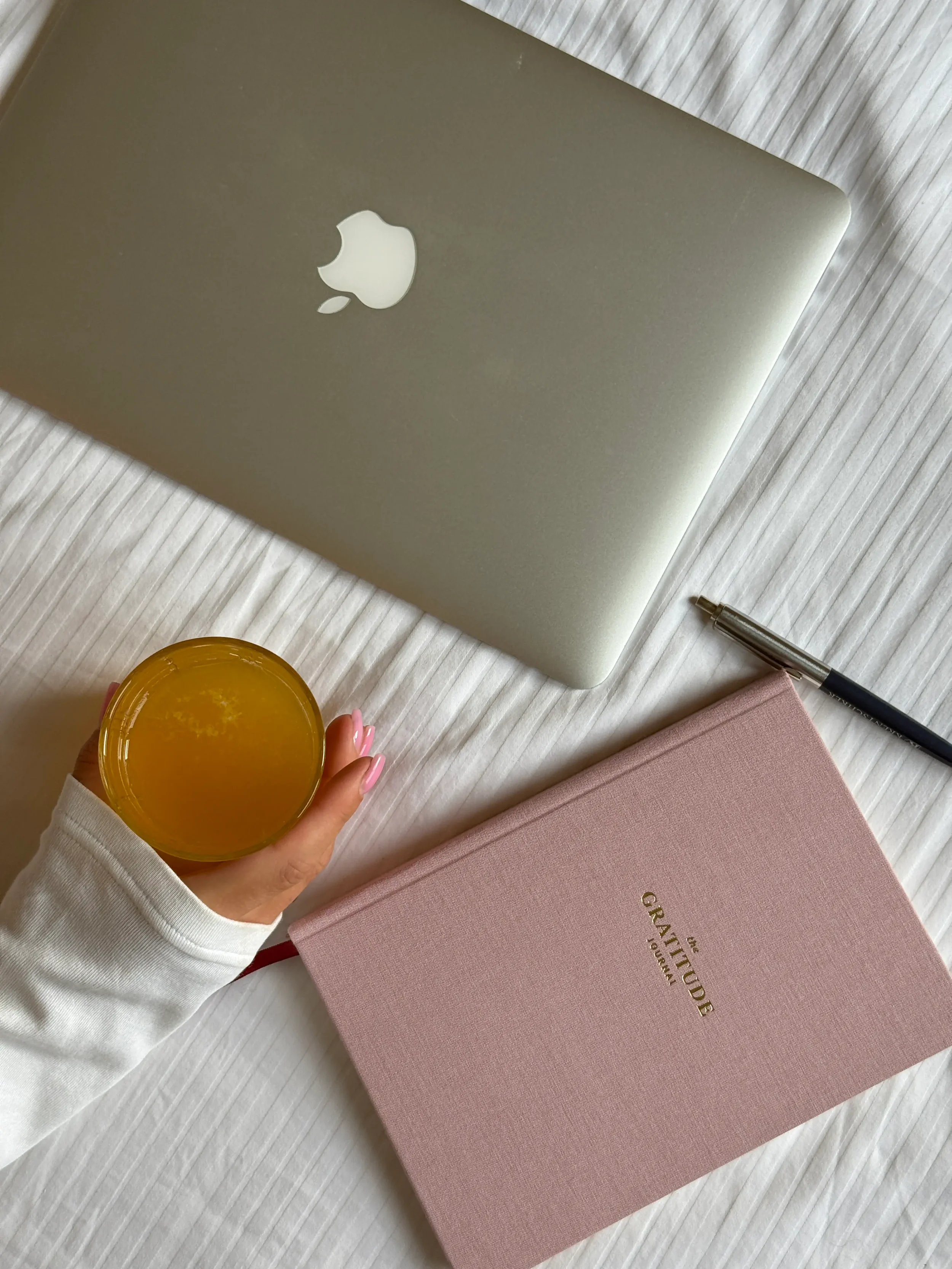 A closed silver MacBook laptop, a pink gratitude journal, a black and silver pen, and a hand holding a glass of orange juice on a white textured surface.