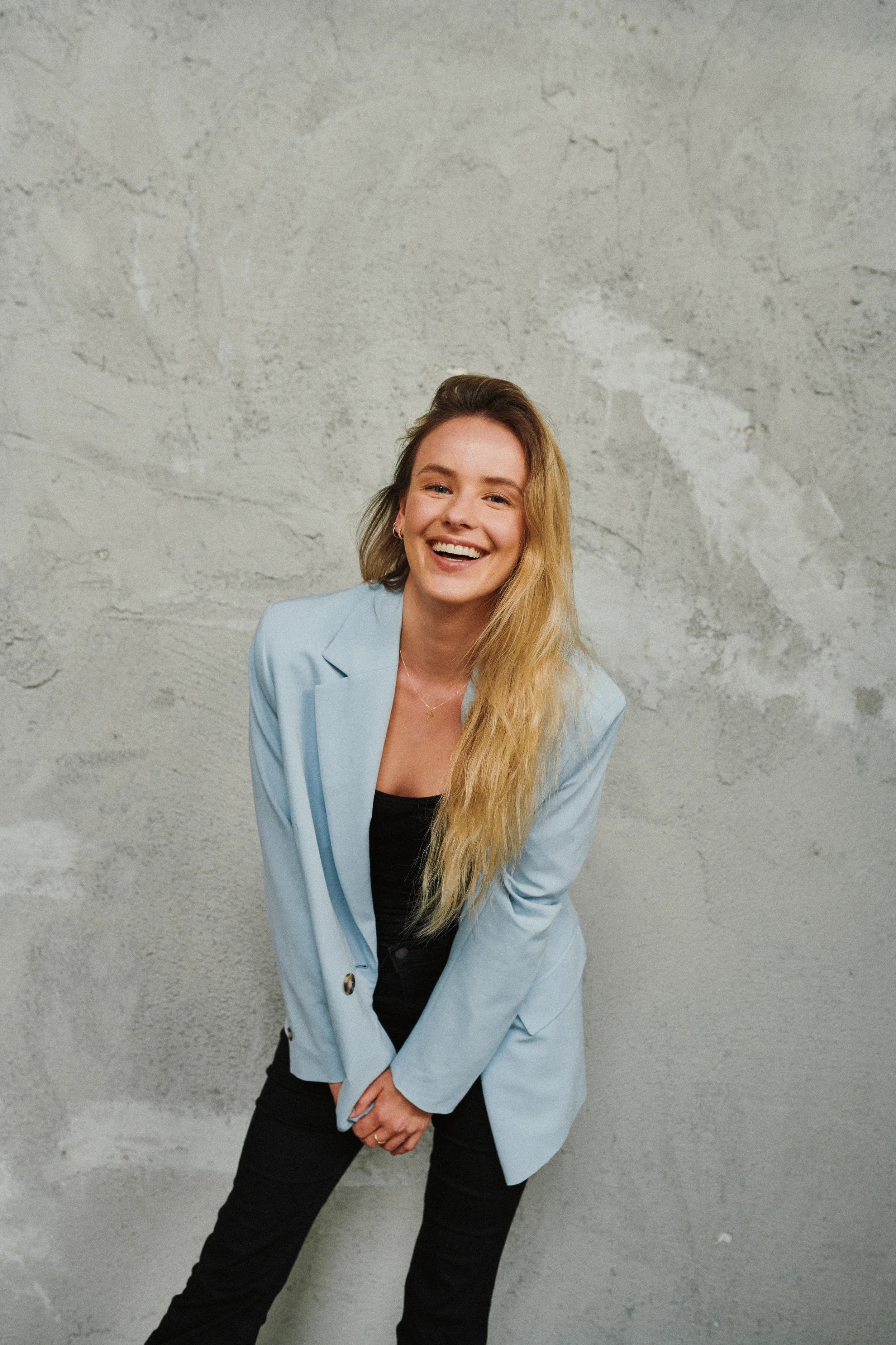 A young woman with long blonde hair smiling while standing against a concrete wall, wearing a light blue blazer over a black top and black pants.