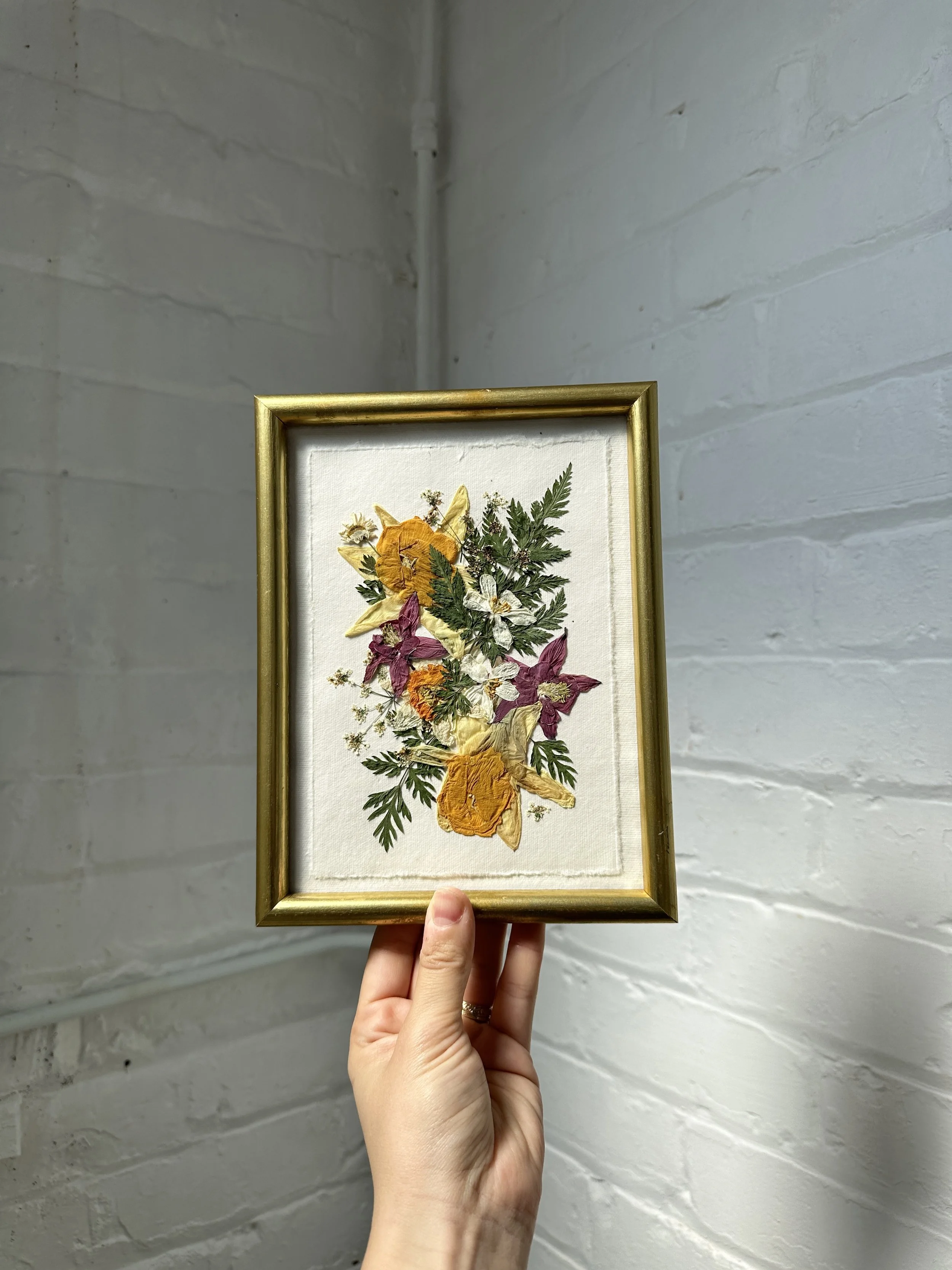Person holding a framed pressed flower art piece featuring yellow, purple, and white flowers with green foliage, against a white brick wall background.