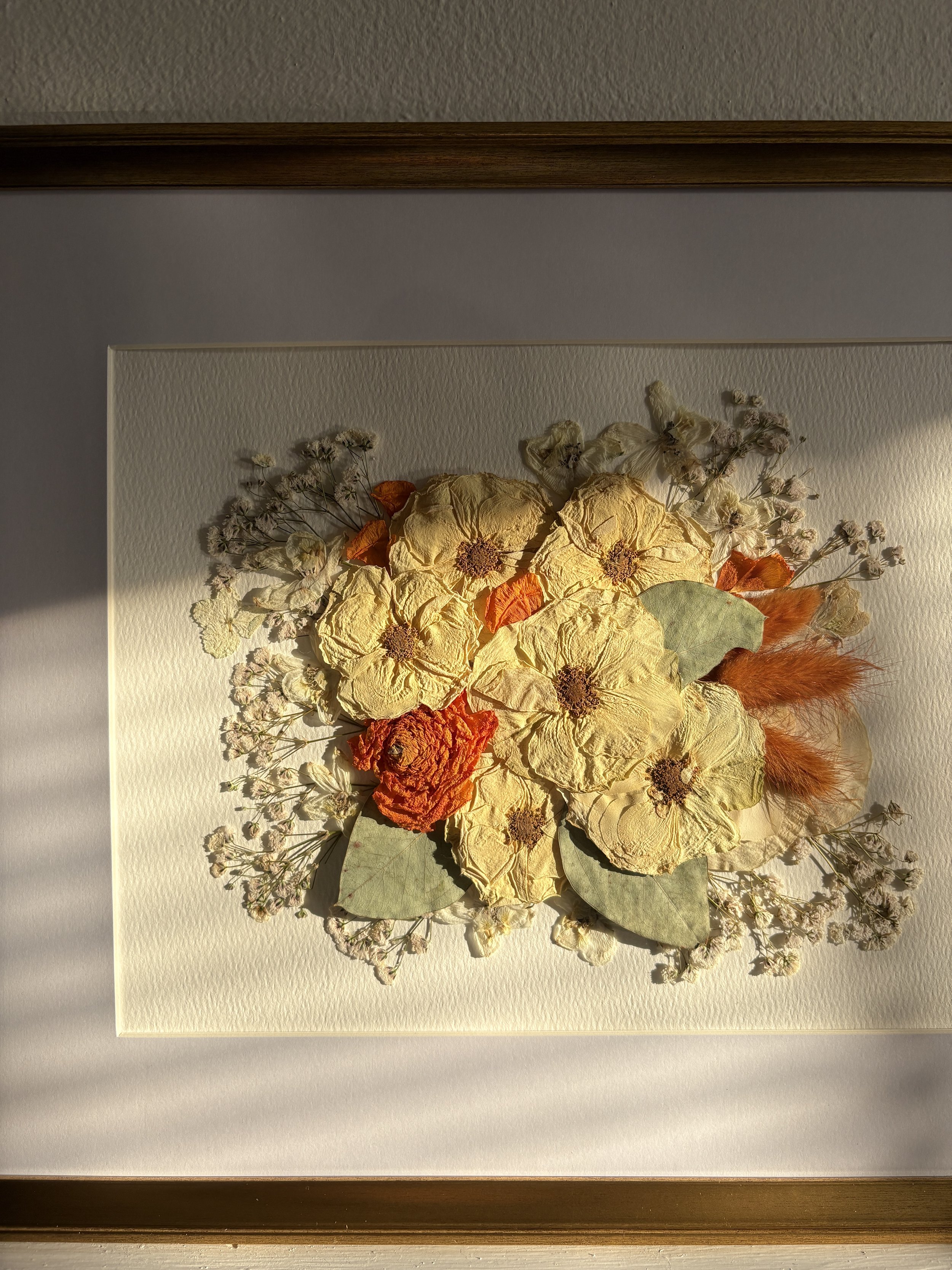Pressed dried flowers arranged as a bouquet with yellow, orange, and cream colors, mounted on a white textured background, framed with wood.