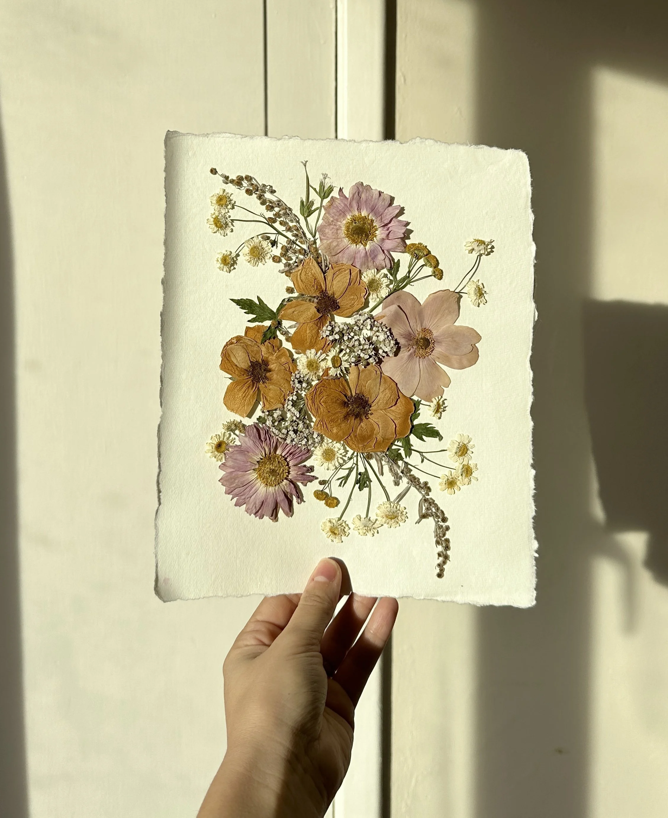 Hand holding a piece of paper with pressed flowers arranged in a floral pattern.