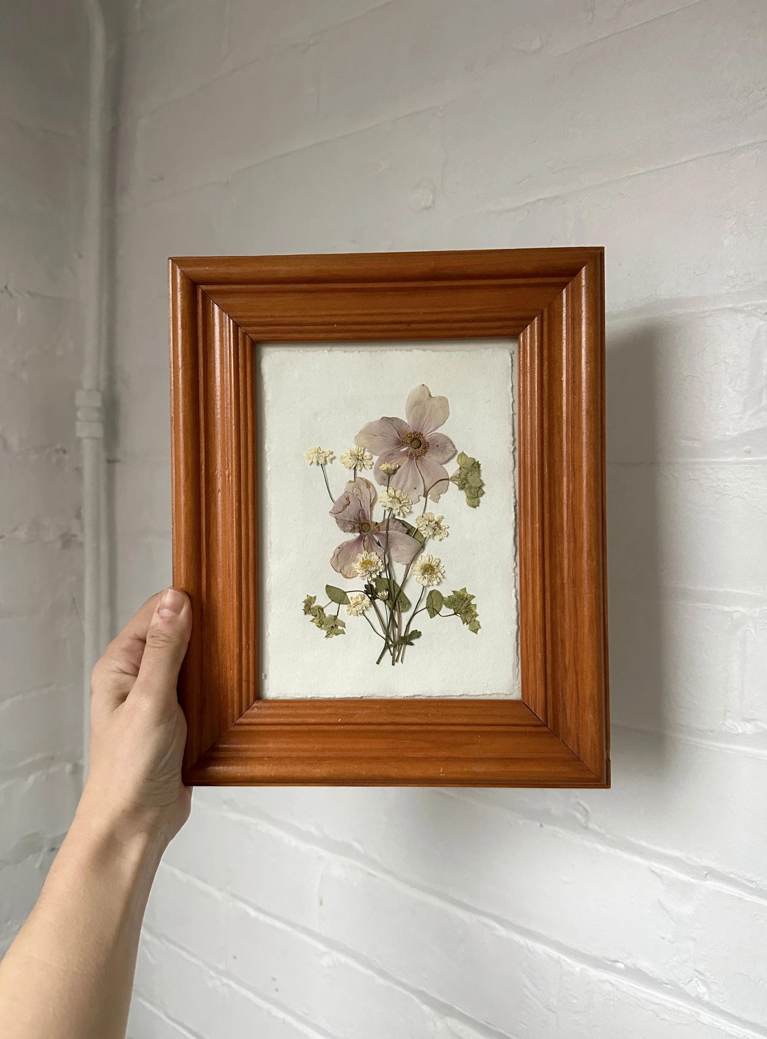 A person holding a framed pressed flower collection against a white brick wall.