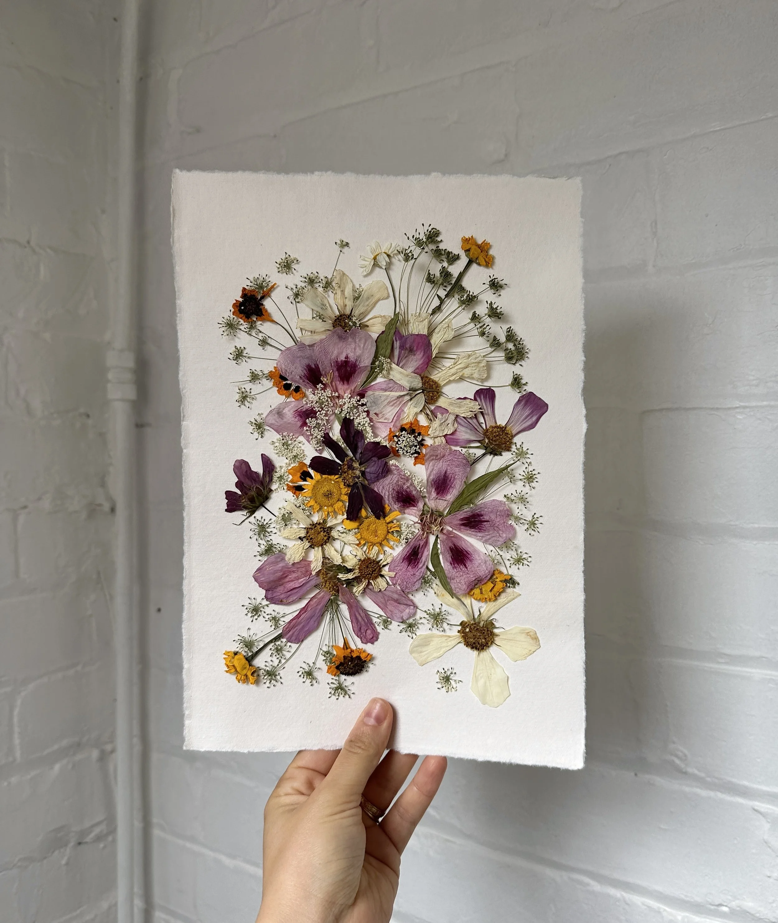 A hand holding a piece of white paper with pressed colorful flowers arranged on it, against a white brick wall background.