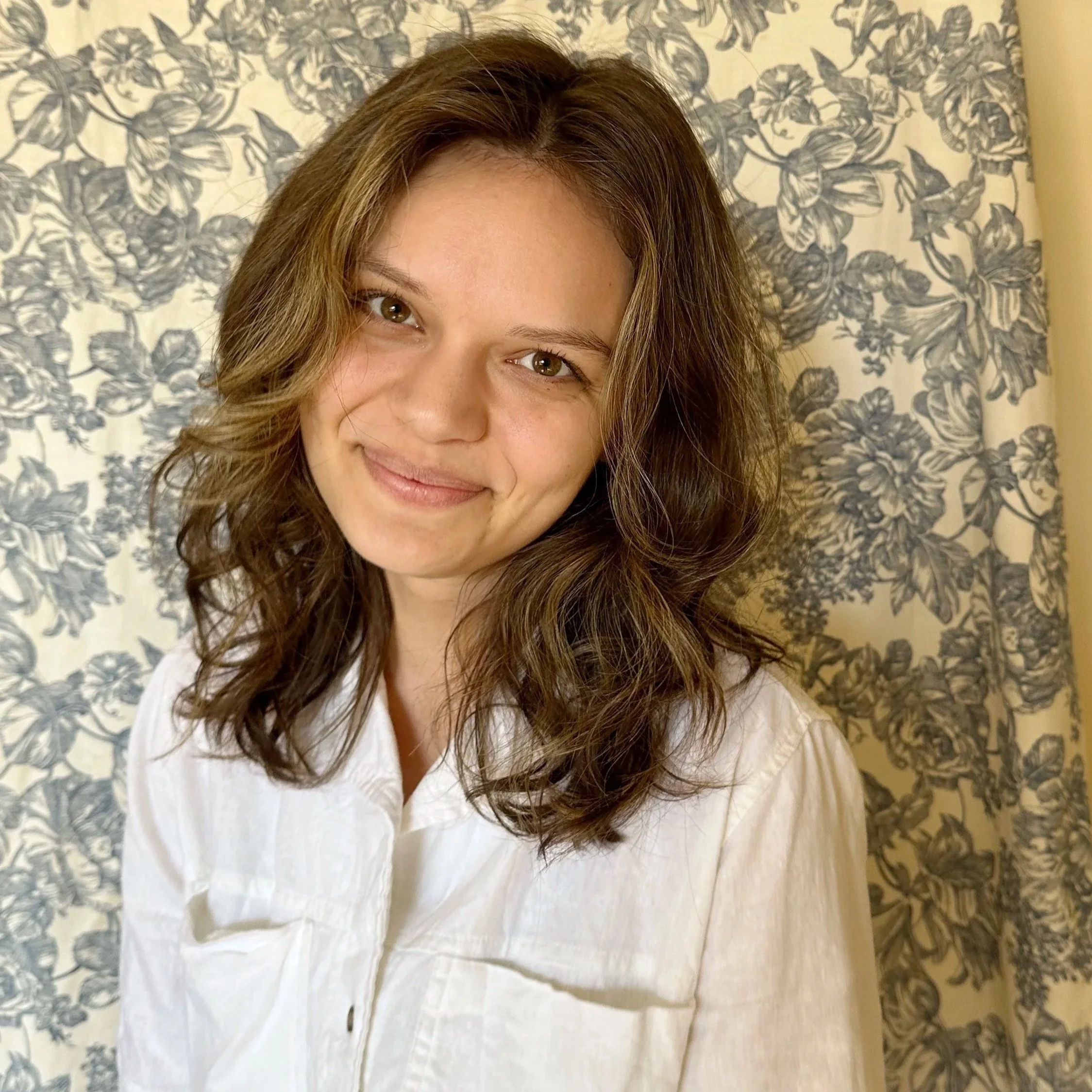 A young woman with wavy brown hair and light skin smiling, wearing a white button-up shirt, standing in front of a floral patterned curtain background.