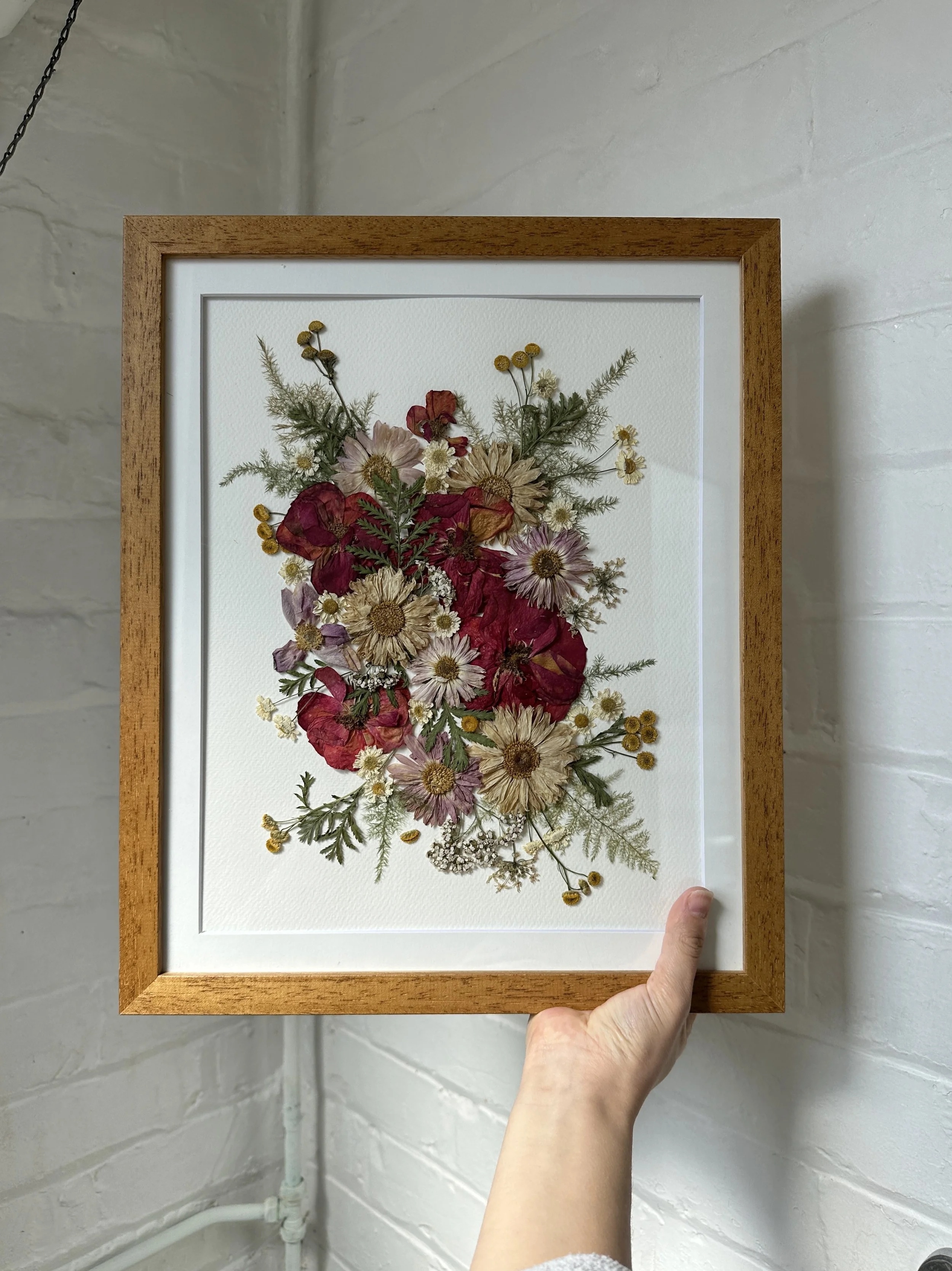Pressed dried flowers arranged in a floral pattern inside a wooden frame, held by a person's hand against a white brick wall.