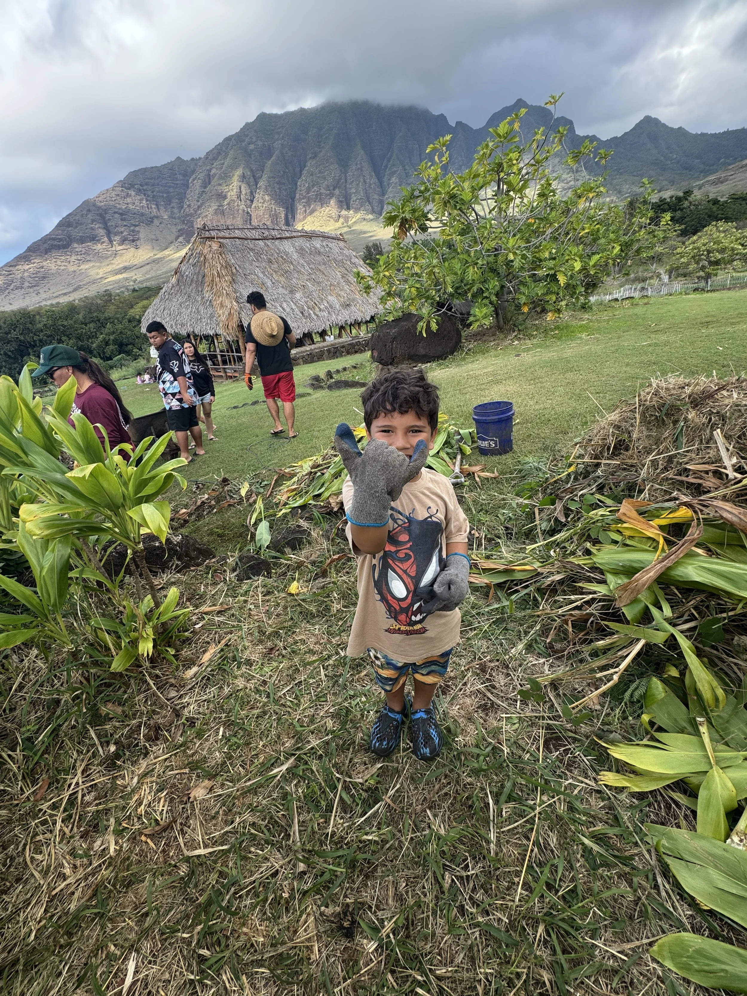 A young boy in a beige Spider-Man t-shirt and striped shorts stands in the foreground, smiling and making a hand gesture. In the background, there are several people near a thatched-roof hut, surrounded by green plants and grass, with mountains and cloudy sky behind.
