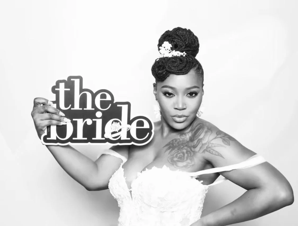 A woman with styled hair and tattoos holding a sign that says "the bride" in front of a plain background.