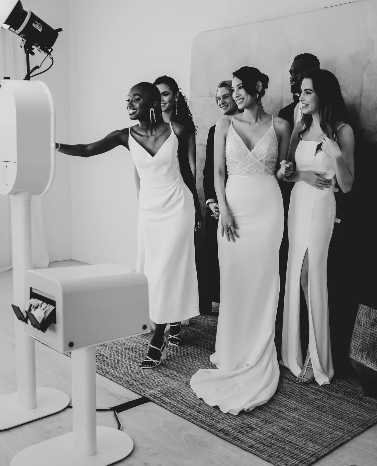 A group of diverse women in elegant white dresses posing for a photo, with one woman adjusting a photo booth camera in a well-lit room.