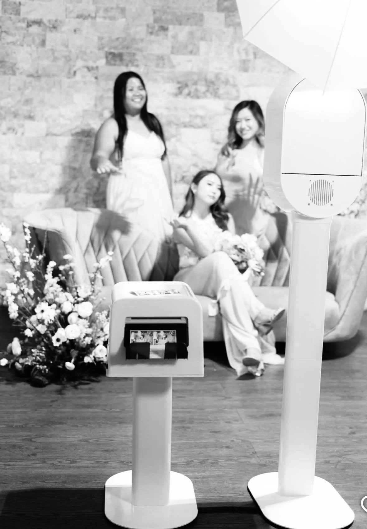Three women in dresses posing for a photo with a bouquet, a photo booth, and a camera in the foreground.