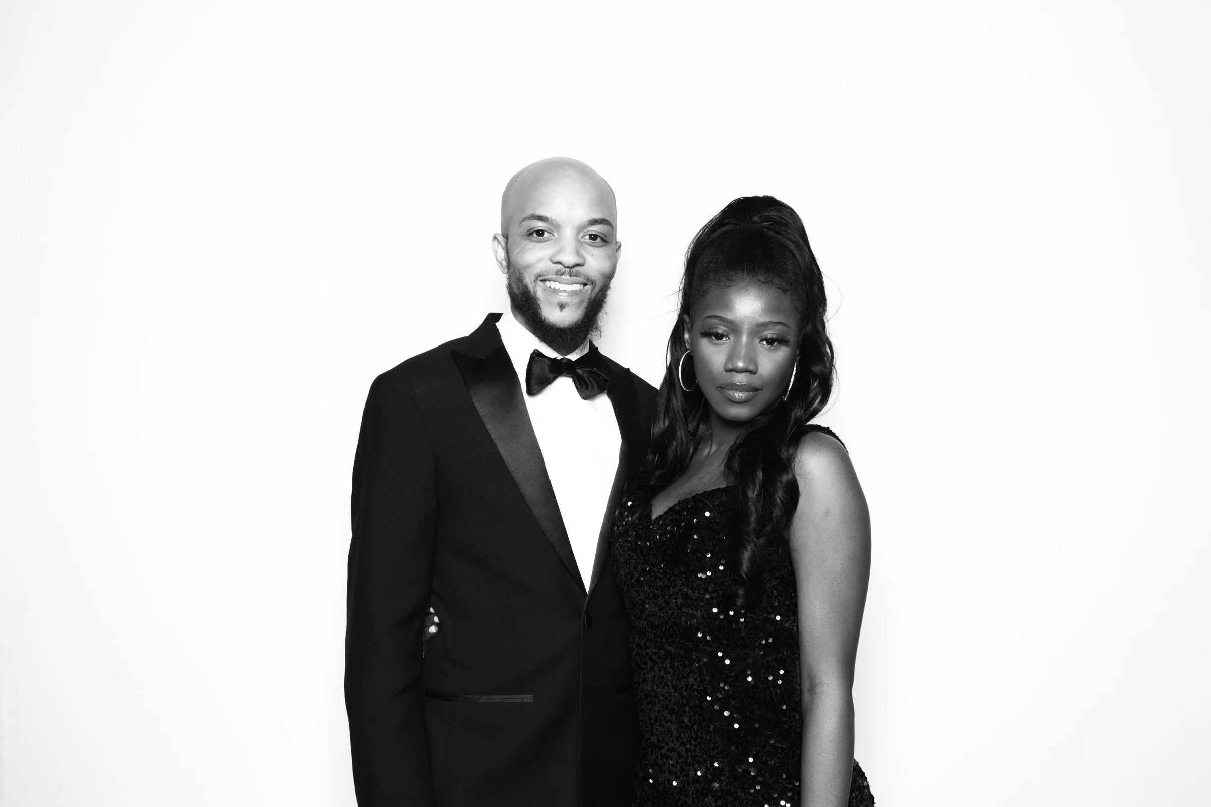 A man in a tuxedo and a woman in a sequined dress posing together against a plain background.