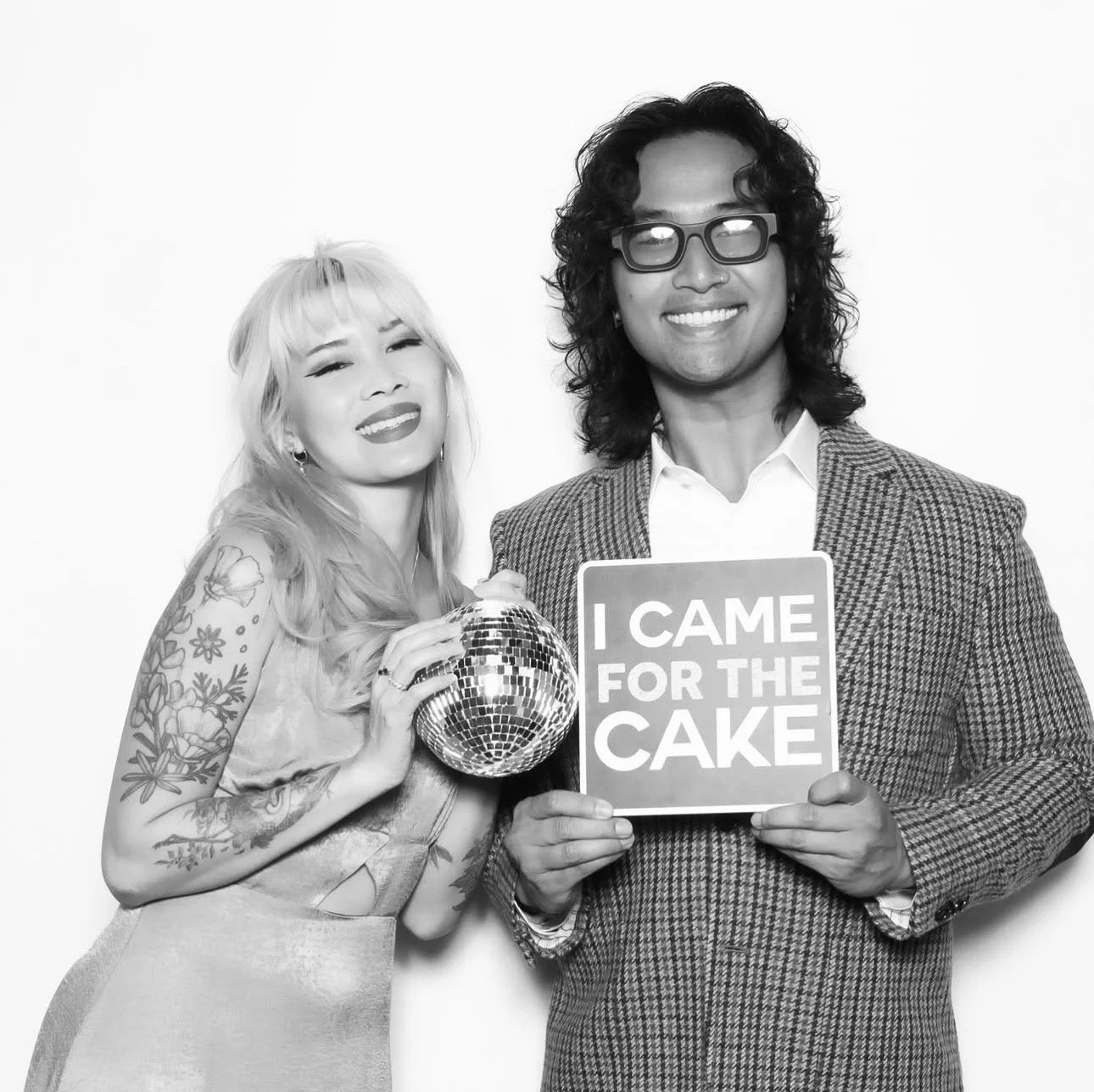 Two people, a woman with tattoos and blonde hair, and a man with glasses and curly hair, standing together against a plain background. The woman is holding a disco ball, and the man is holding a sign that says "I CAME FOR THE CAKE." Both are smiling.