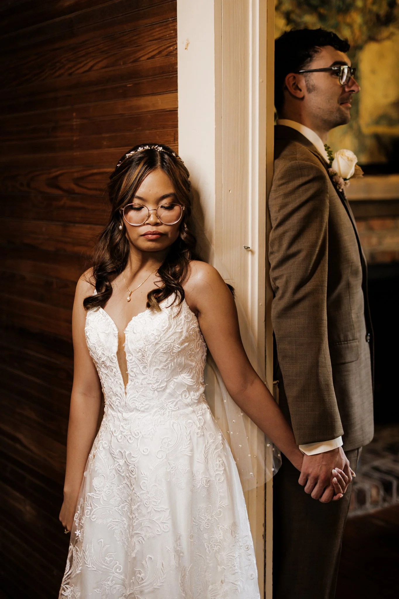 A bride and groom standing back-to-back, holding hands, with their backs against a wall. The bride is wearing a white lace wedding dress and glasses, and has long wavy hair. The groom is wearing a brown suit, glasses, and has short dark hair. They are standing indoors, with a wooden wall behind the bride and a blurred background behind the groom.