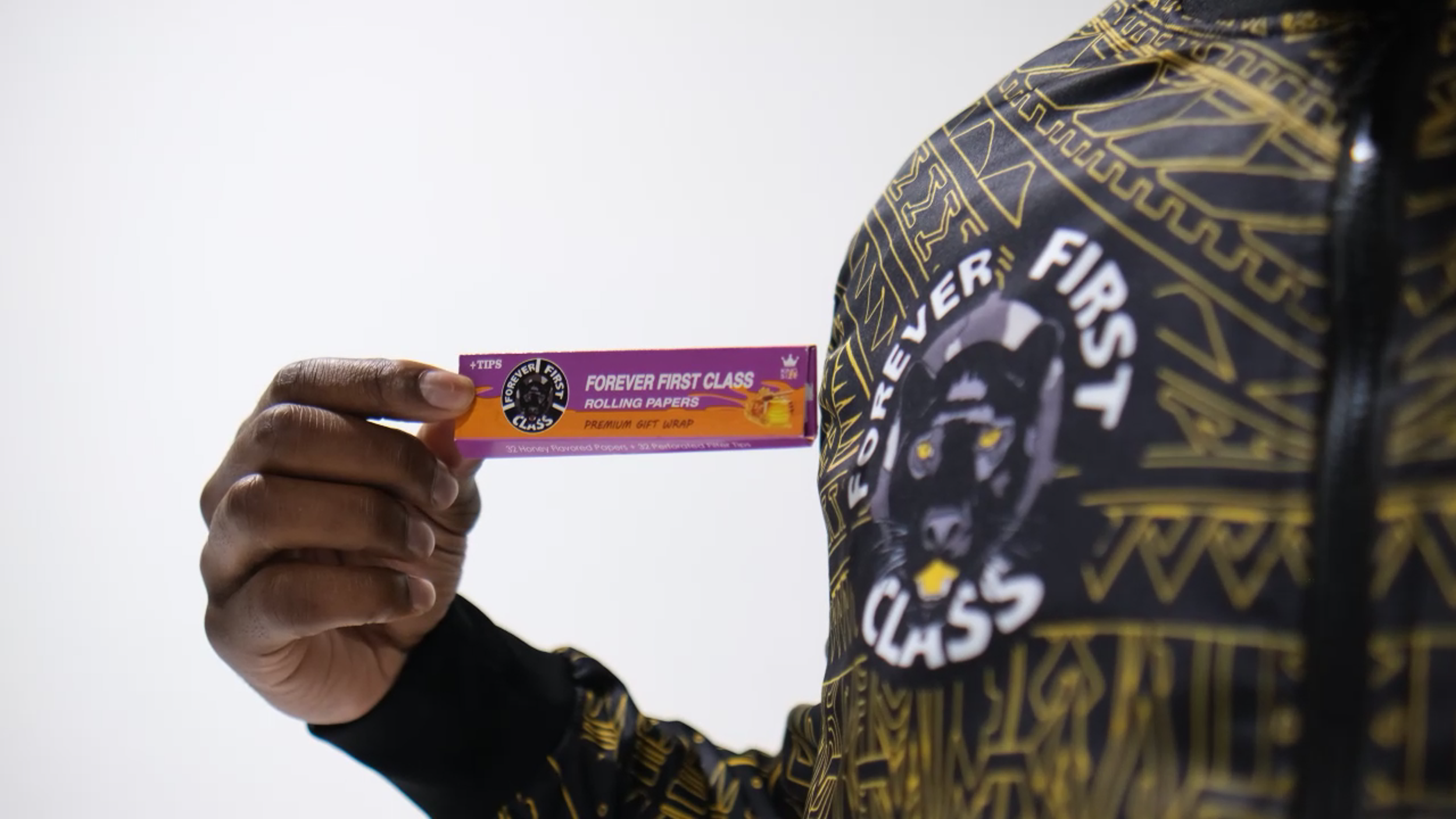 Close-up of a person holding a pack of Forever First Class rolling papers next to their chest, wearing a black and gold patterned hoodie with a lion and text logo.