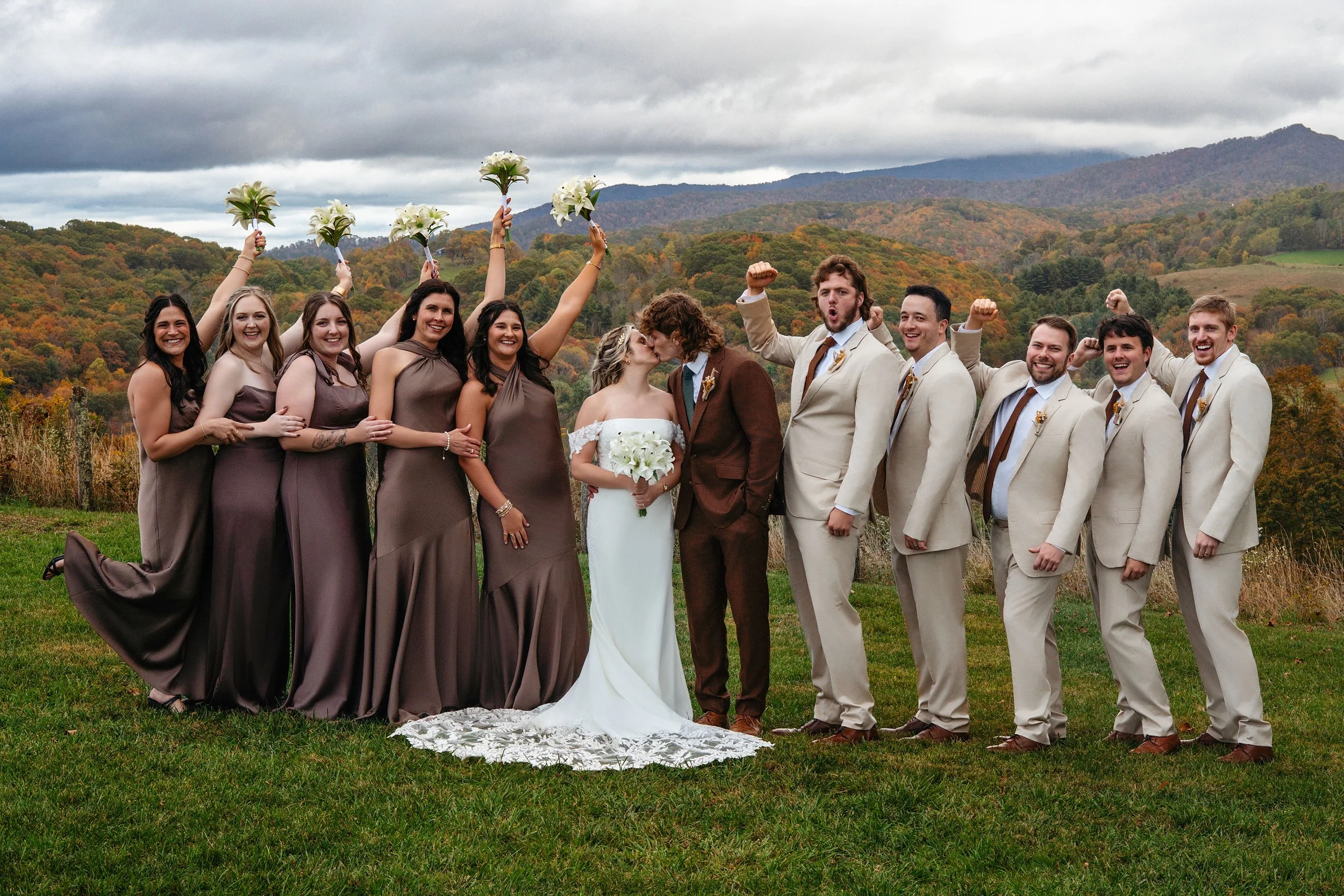 A wedding party stands outdoors in a field with a backdrop of colorful fall foliage and mountains. The bride and groom kiss in the center, surrounded by bridesmaids holding bouquets and groomsmen raising fists in celebration.