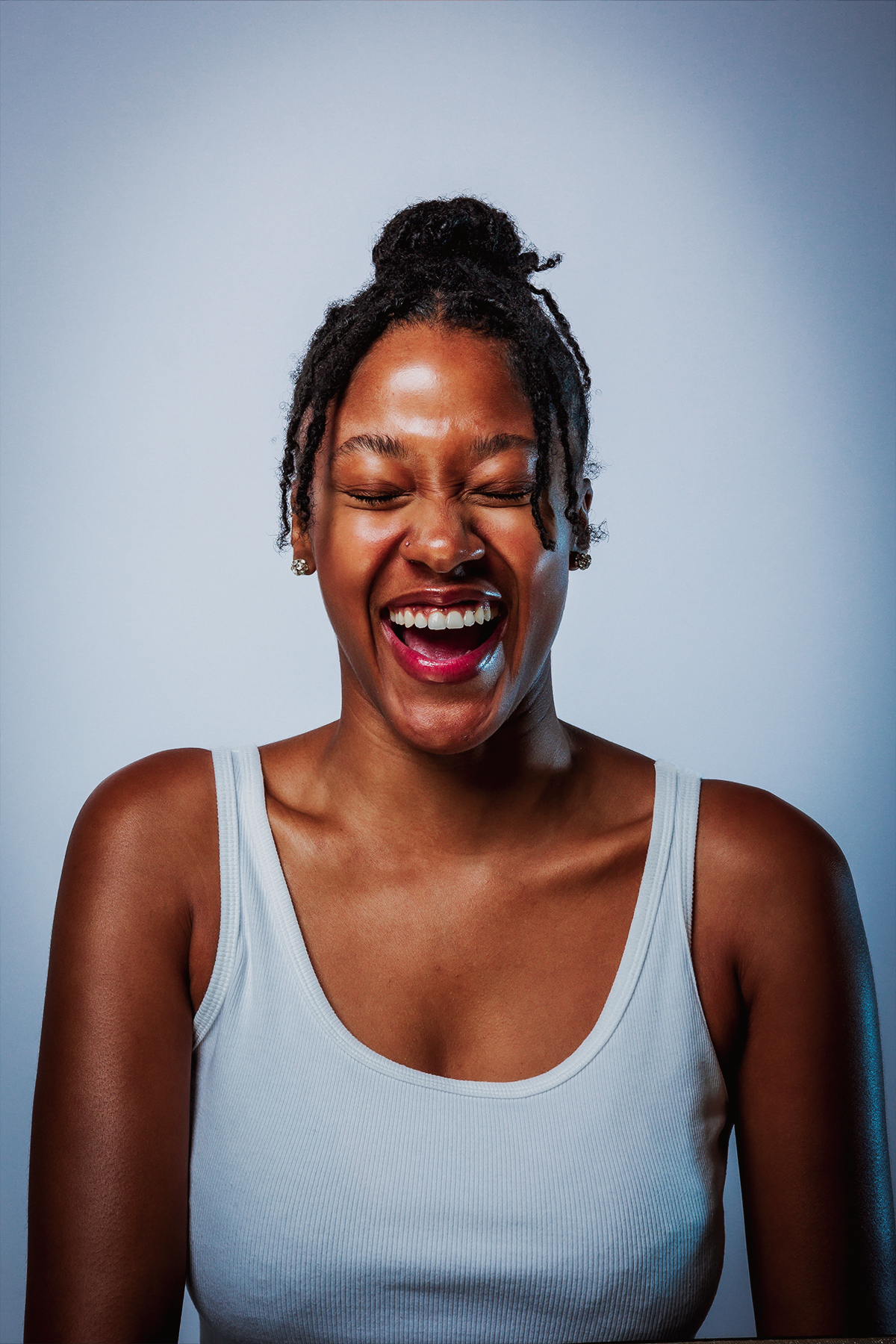 A woman with dark skin and dreadlocks tied up in a bun laughing with her eyes closed, wearing a white tank top against a plain gray background.