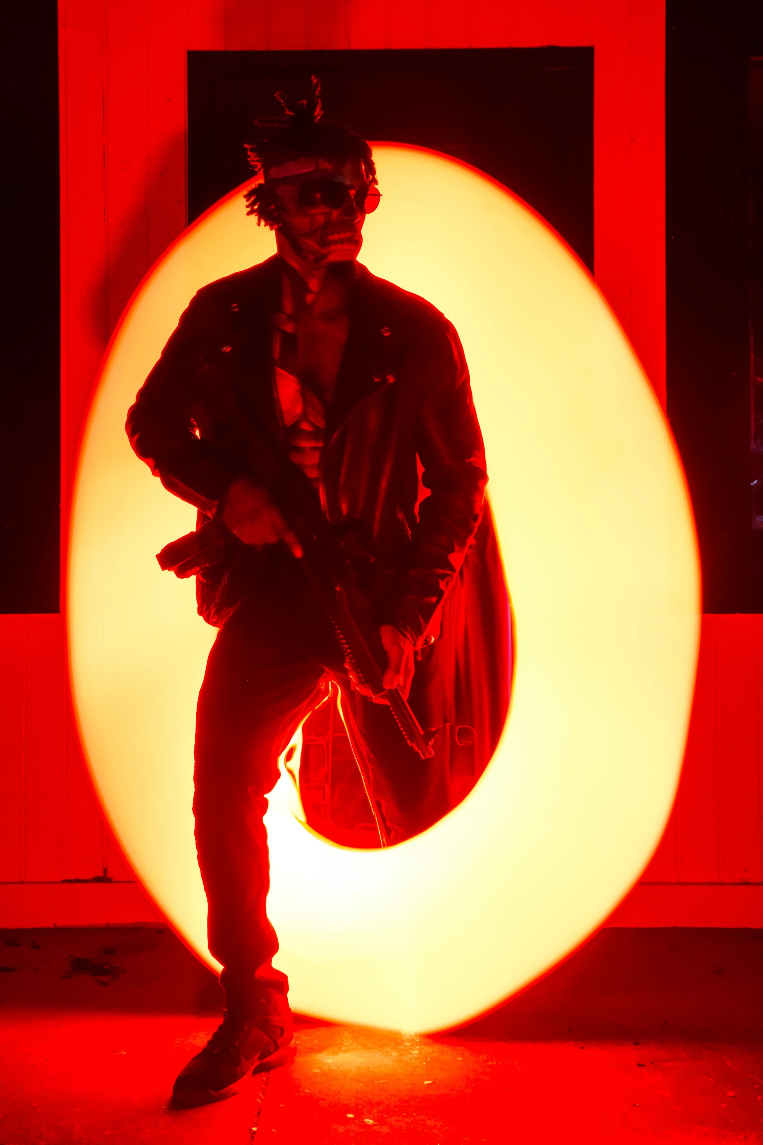 A person wearing a leather jacket, striped shirt, and sunglasses, holding a gun, standing in front of a large illuminated orange circle, in a red-lit room.