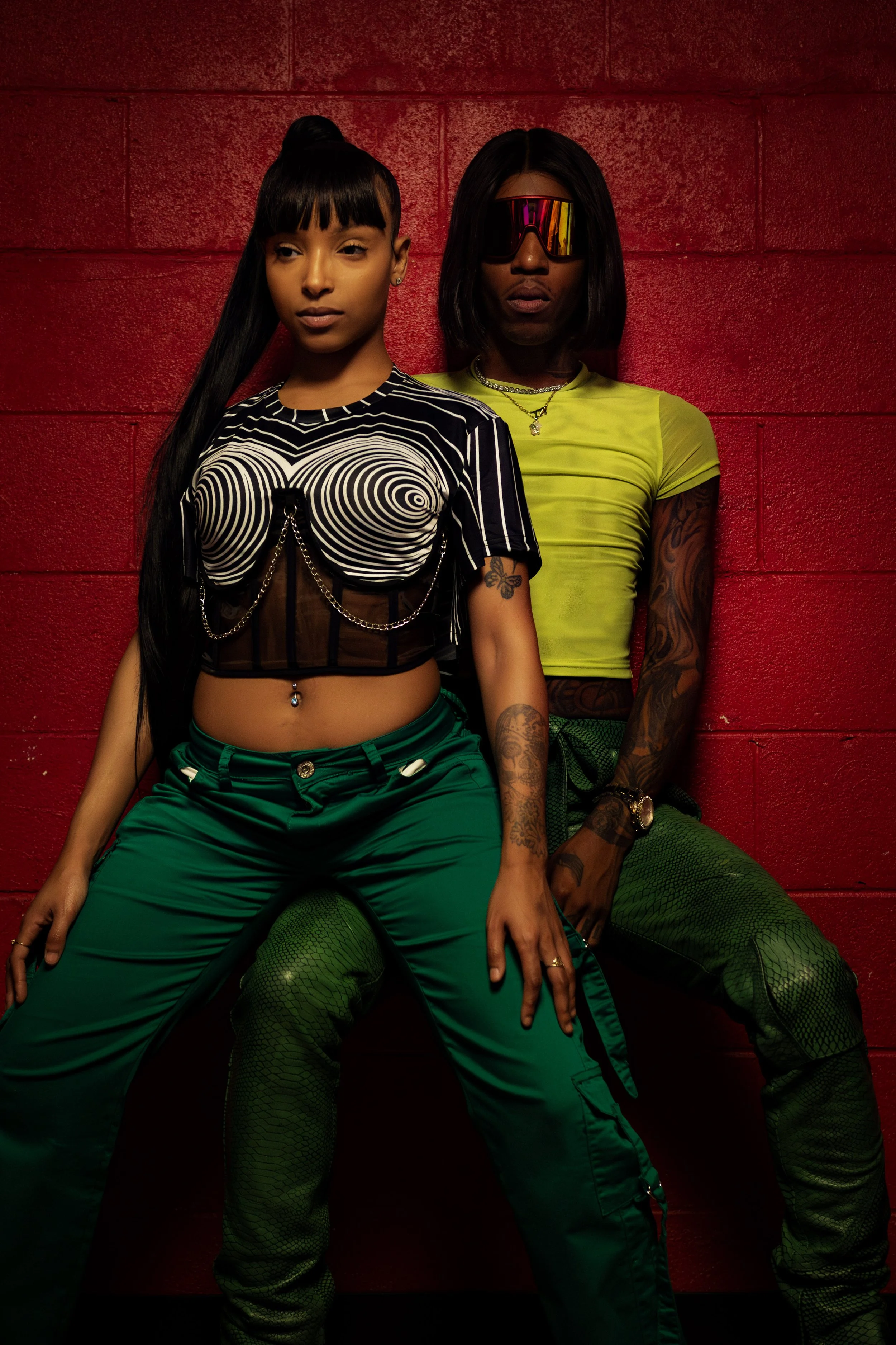 Two fashionable women posing against a red wall; one with long black hair and the other wearing large reflective sunglasses; both dressed in trendy, colorful outfits with tattoos and accessories.