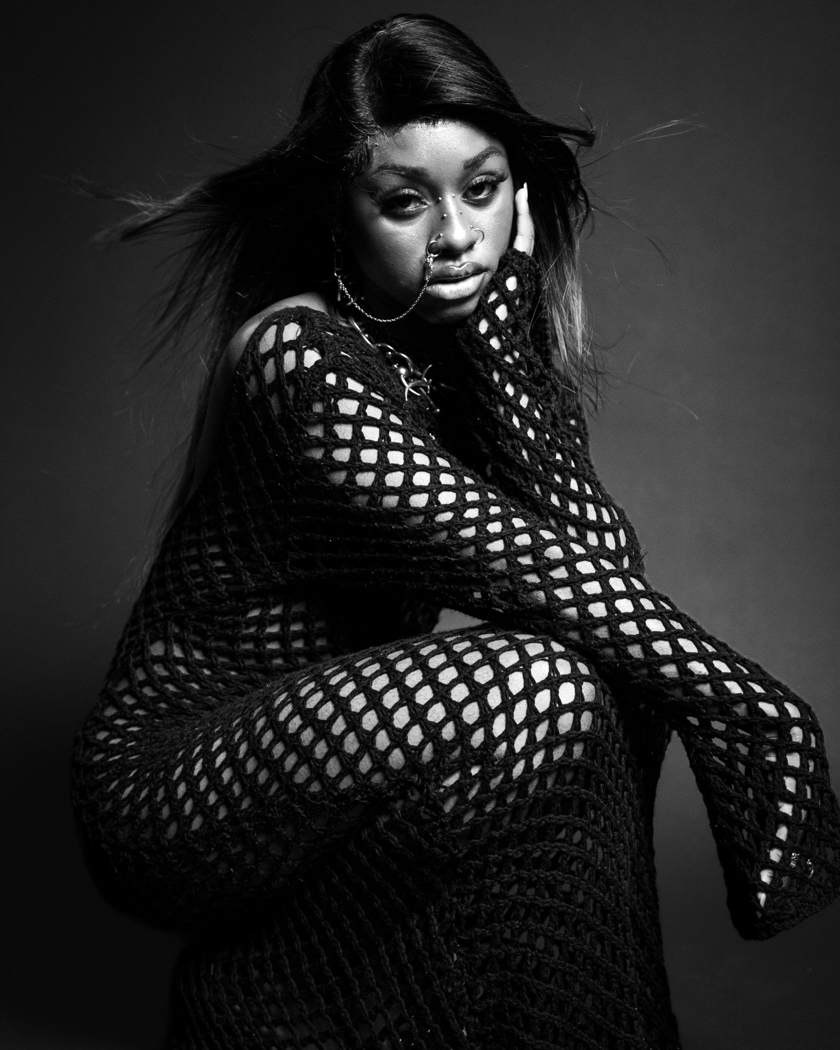 A woman with long hair wearing a black fishnet dress, with multiple facial piercings and jewelry, in a black and white studio portrait.