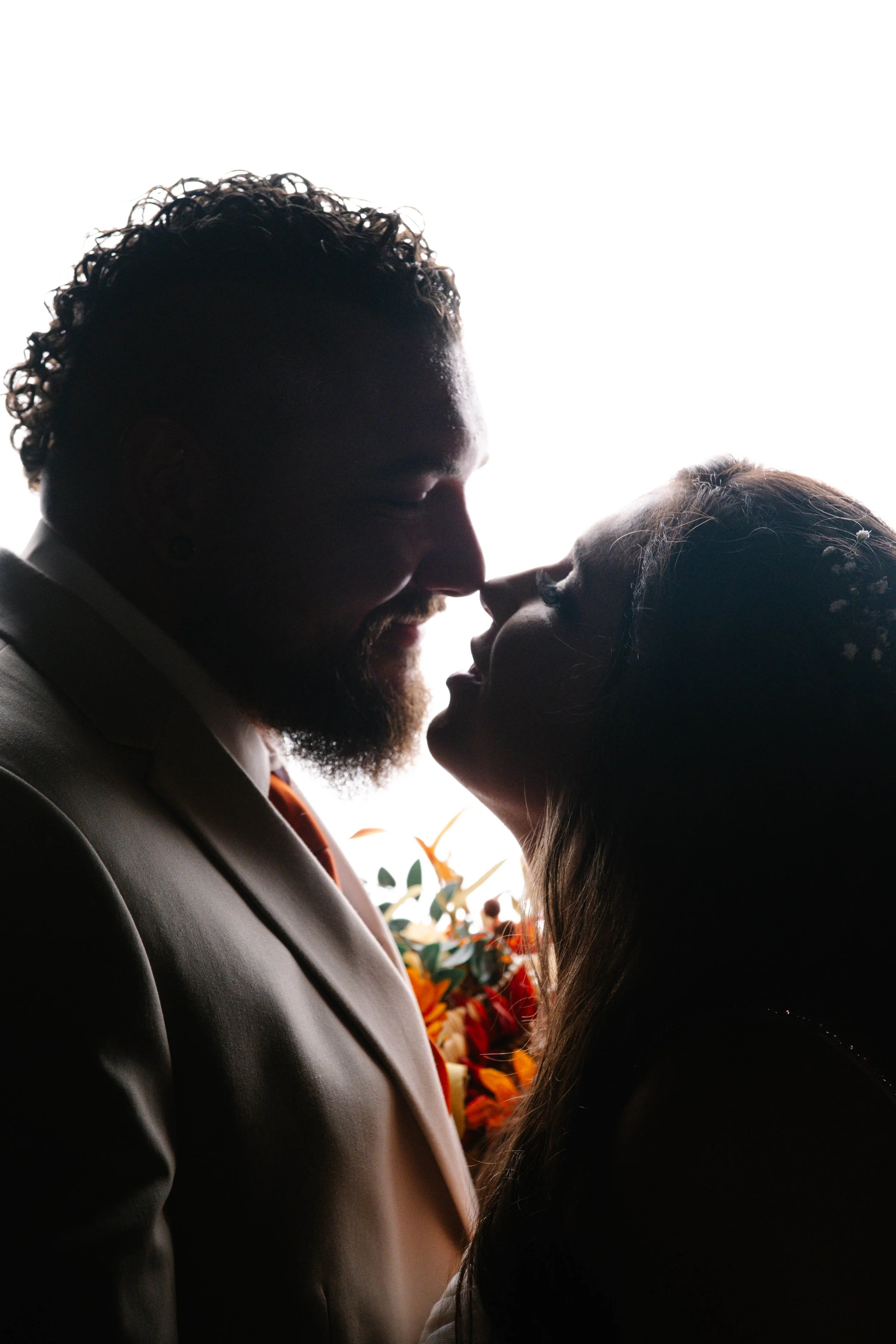Silhouetted couple with noses touching, smiling, with a bouquet of flowers in the background.