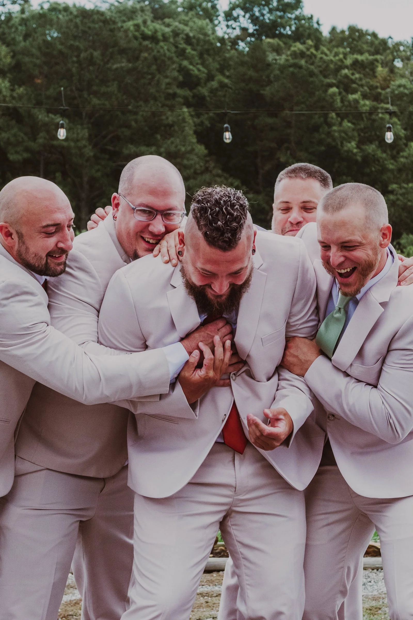 Six men dressed in light-colored suits, engaged in a playful, joyful moment outdoors, surrounded by greenery and hanging string lights.