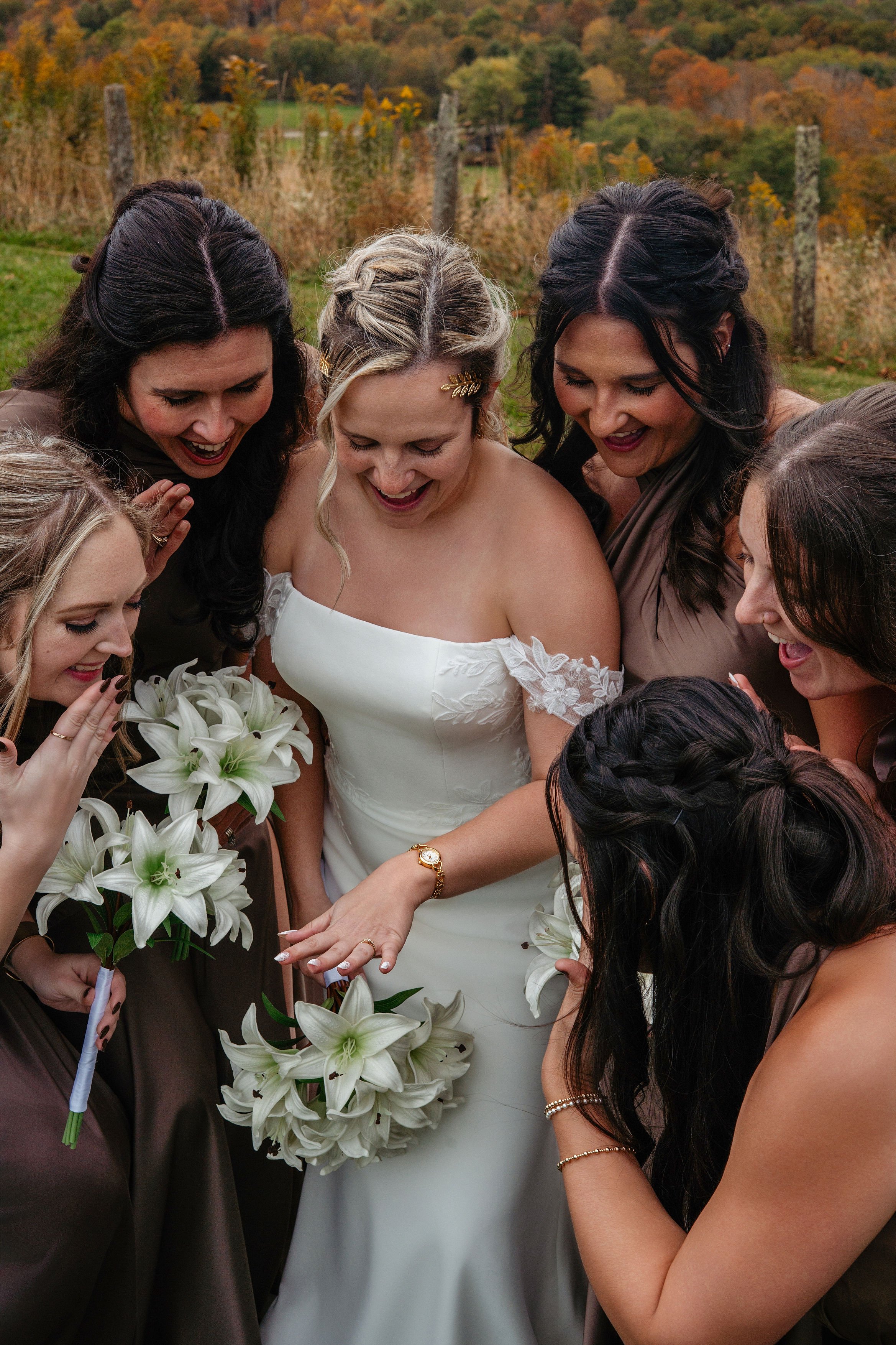 A bride showing off her wedding ring surrounded by her bridesmaids in an outdoor setting with fall foliage.