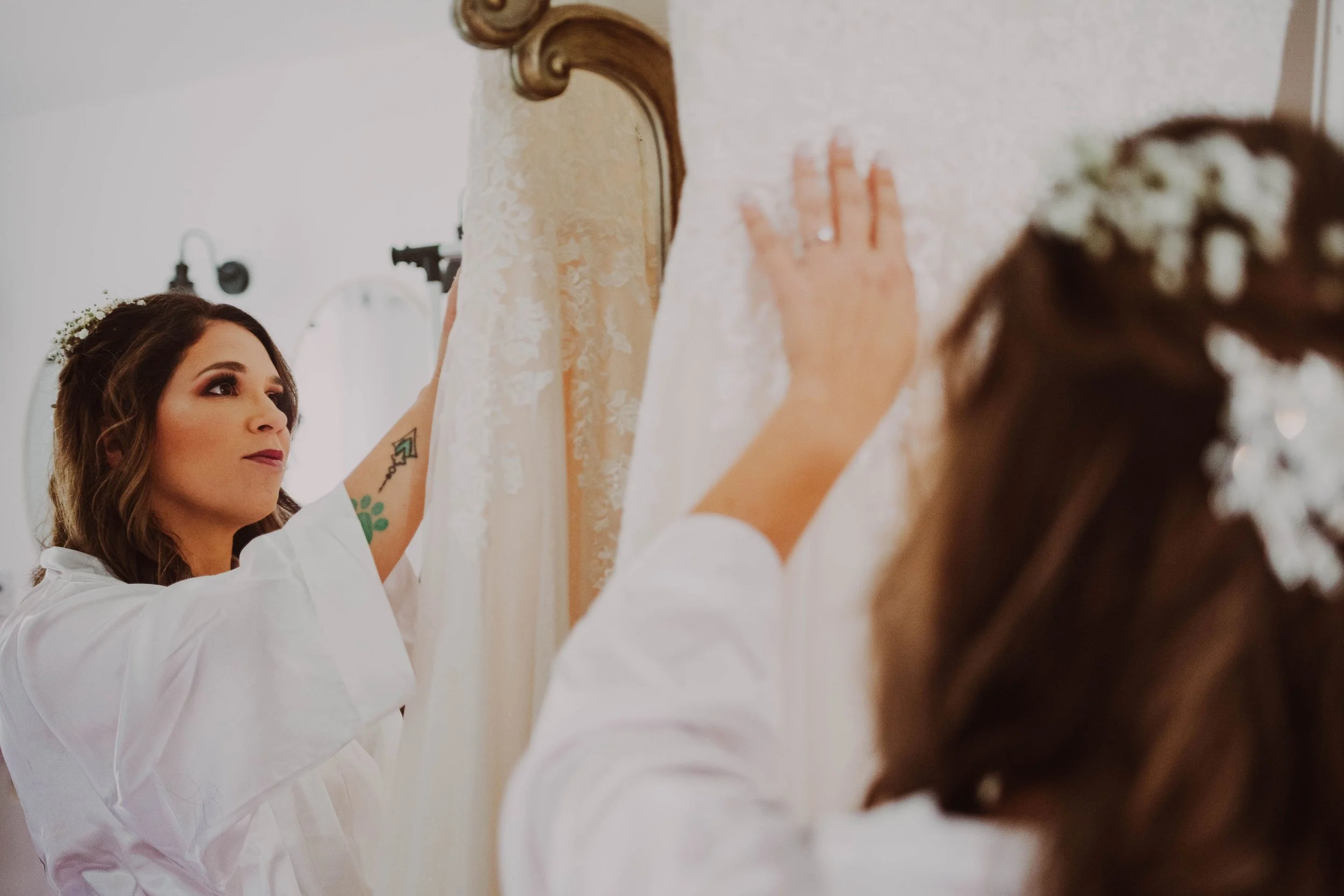 Bridal preparation scene, woman gently touching her wedding dress on a vintage mirror, with hair adorned with white flowers.