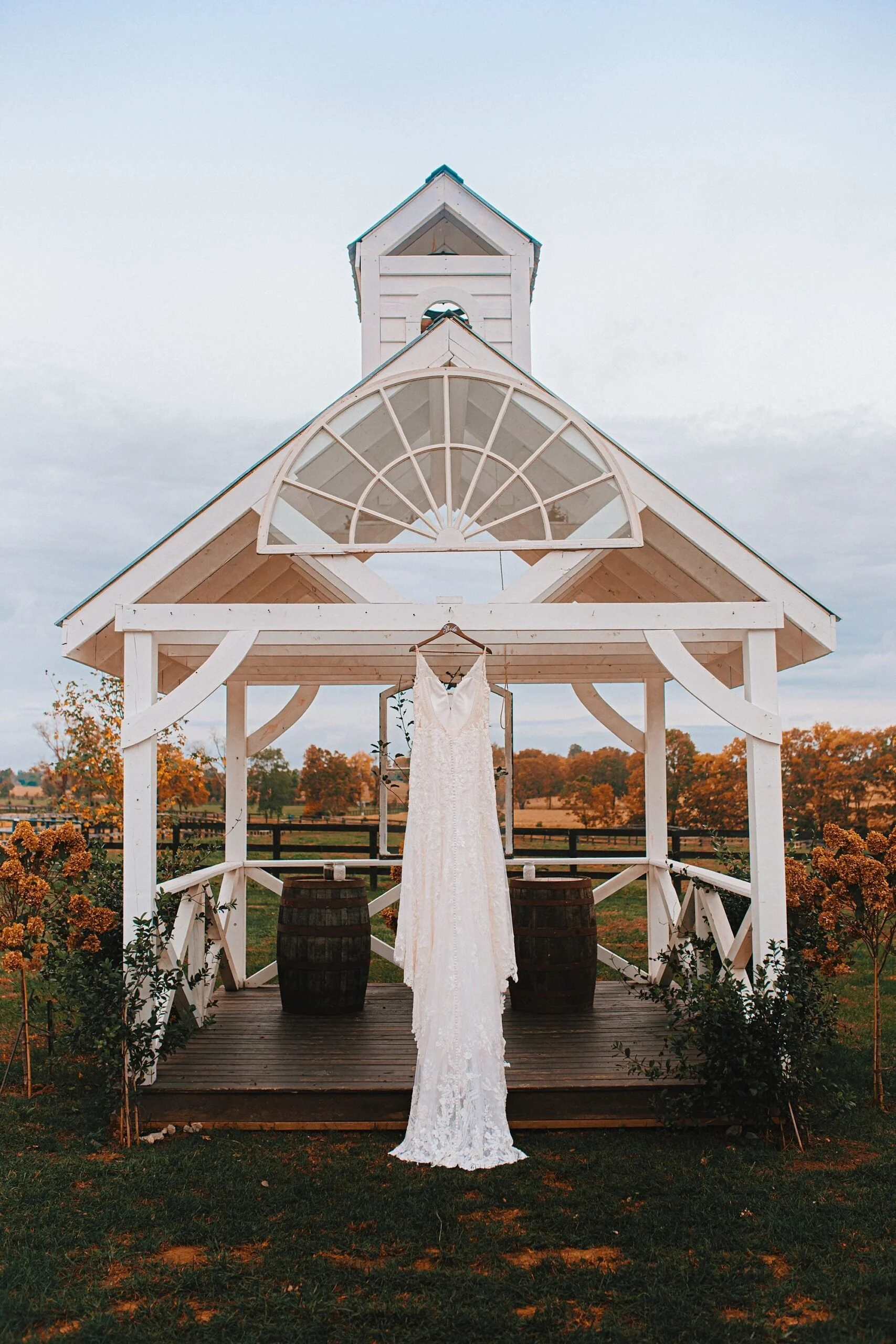 A wedding dress hanging in front of a white wooden gazebo on a farm with fall foliage in the background.