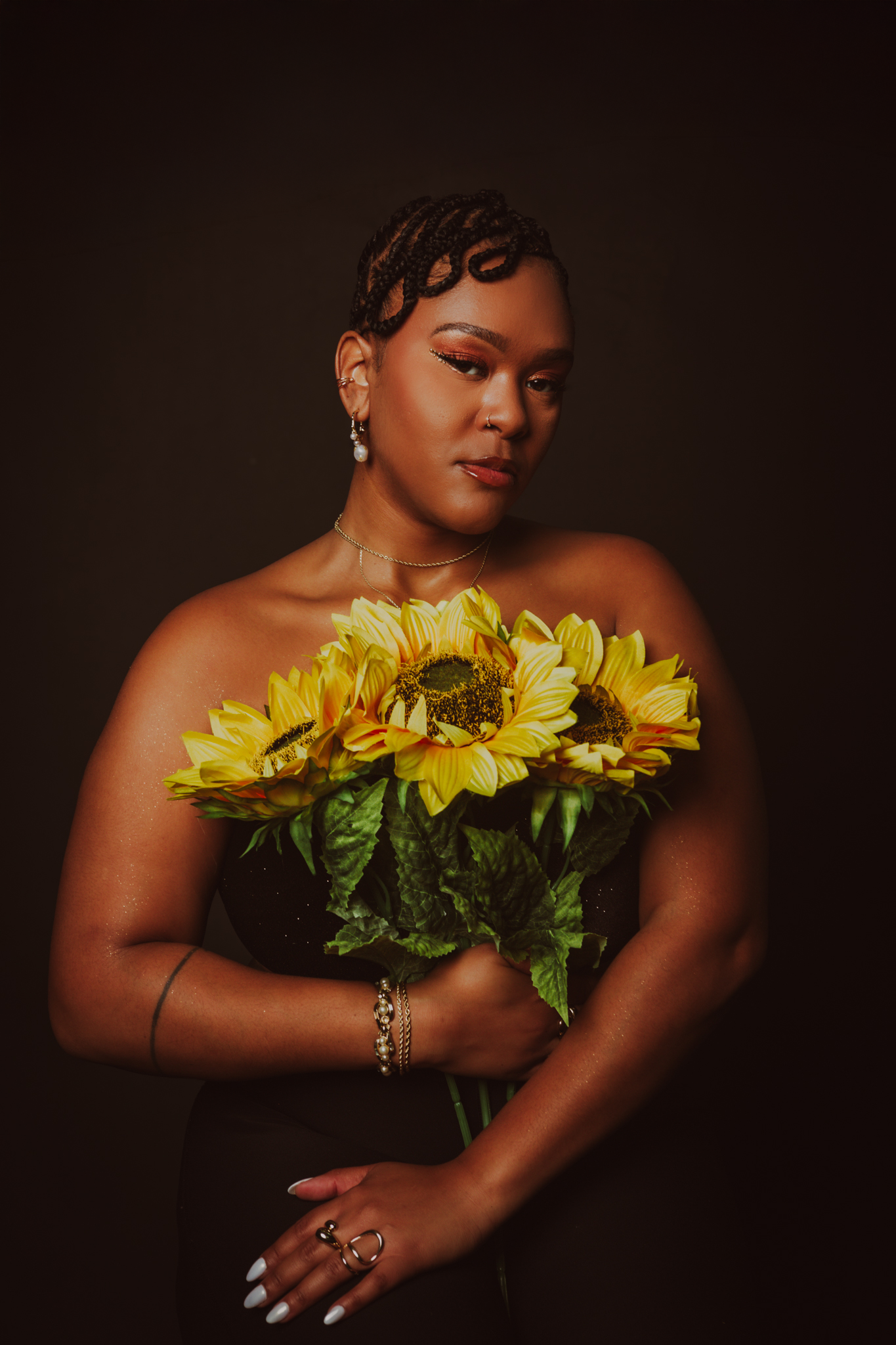 A woman with braided hair holding a bouquet of yellow sunflowers, wearing jewelry against a dark background.