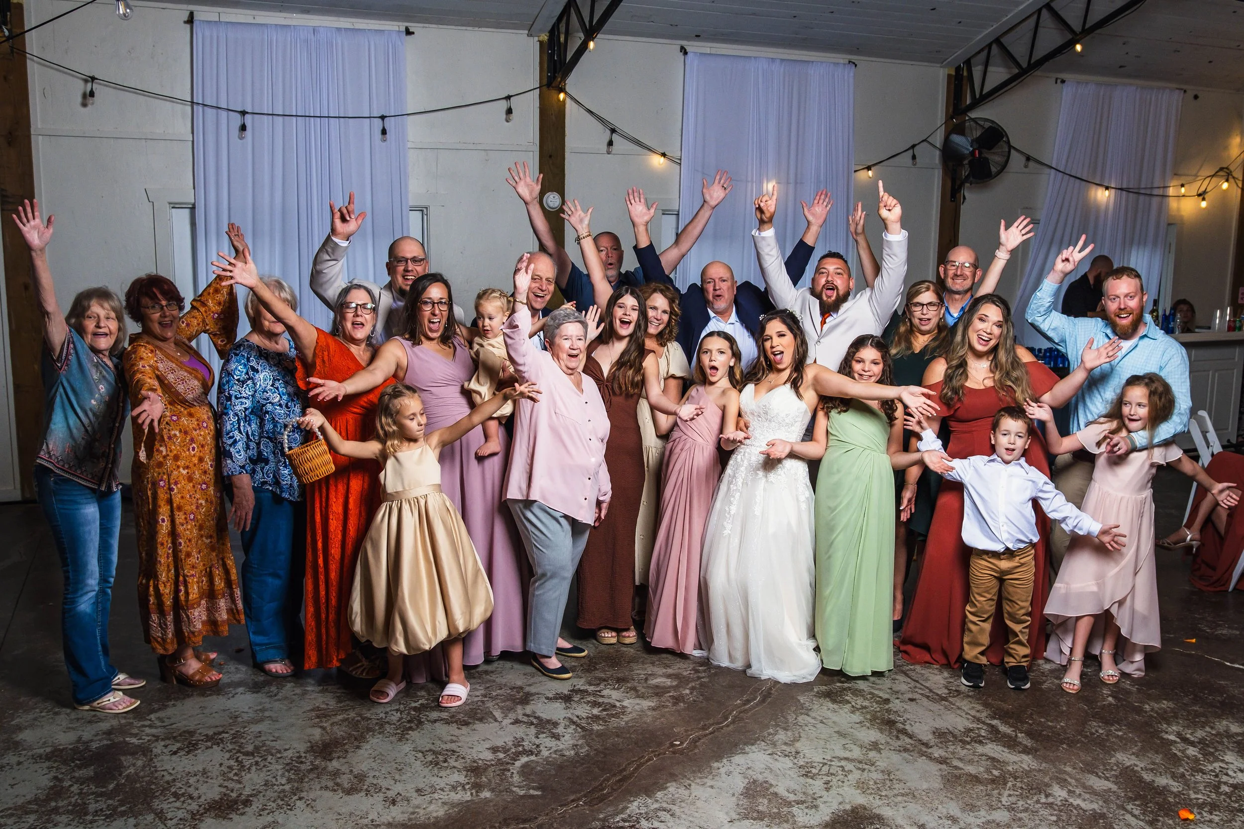 Group of people celebrating at a wedding reception, posing for a photo with smiles and raised hands, indoors with string lights and white curtains.