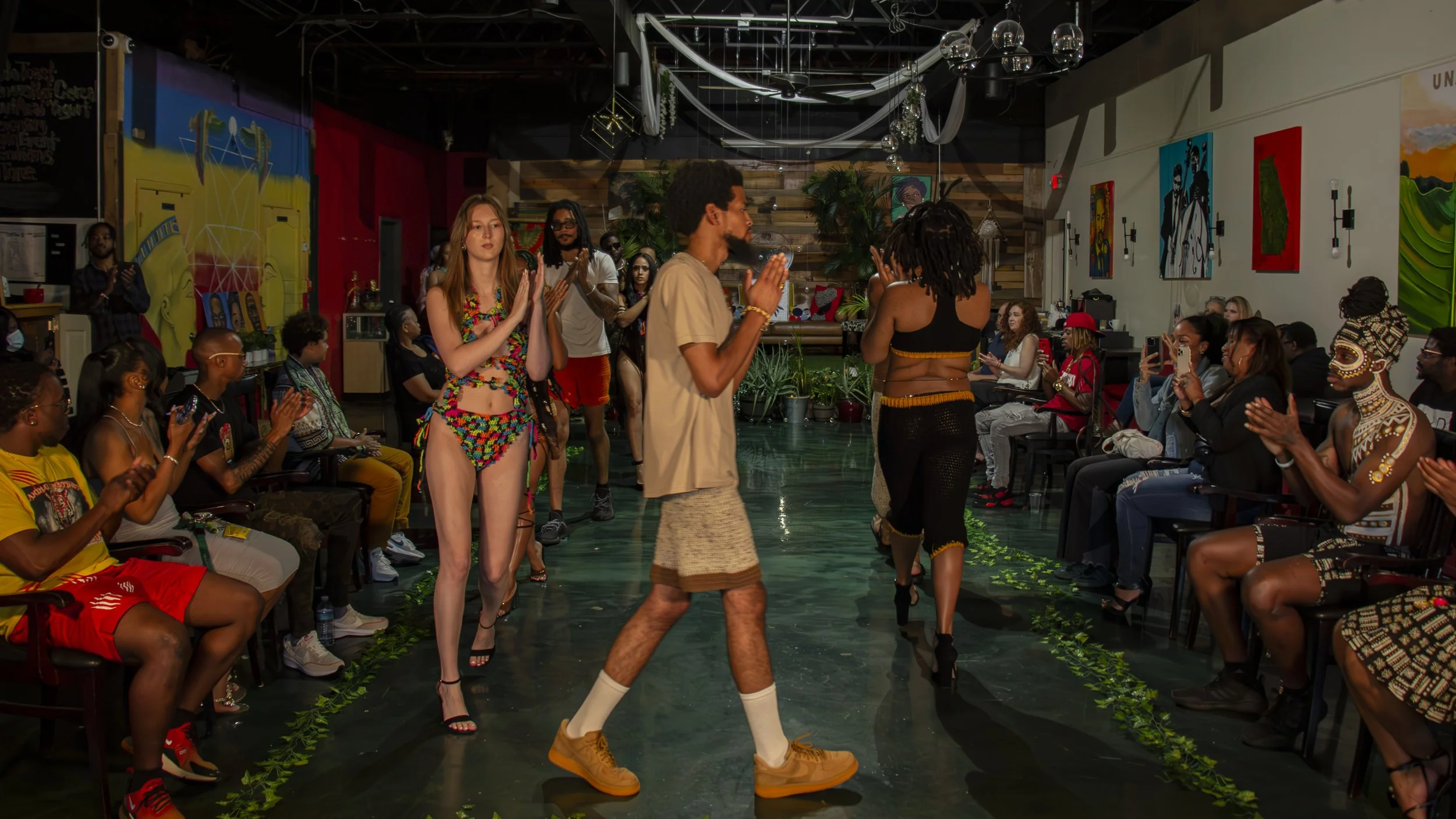 Models walk the runway in colorful, vibrant outfits at a fashion show, with an audience clapping and taking pictures in a decorated indoor venue.