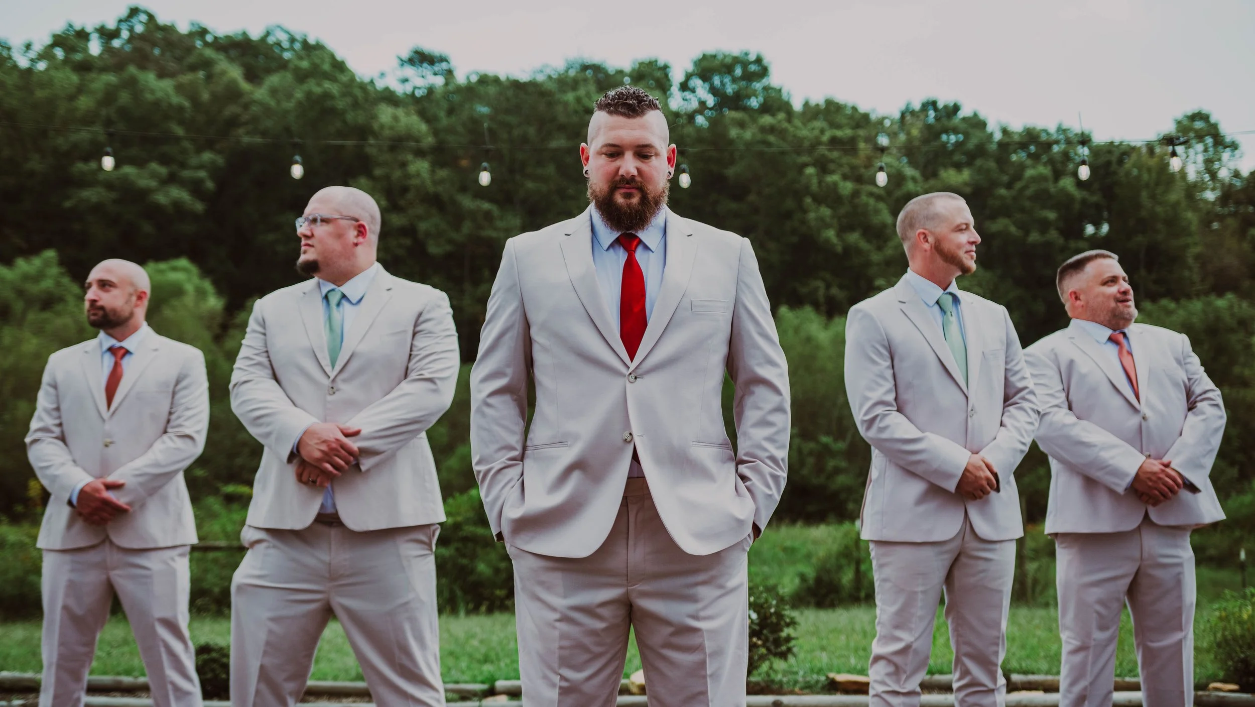 Five men dressed in white suits with colorful ties standing outdoors in front of green trees, with string lights above them. They are standing in a line, with the man in the center having his hands in his pockets and looking down.