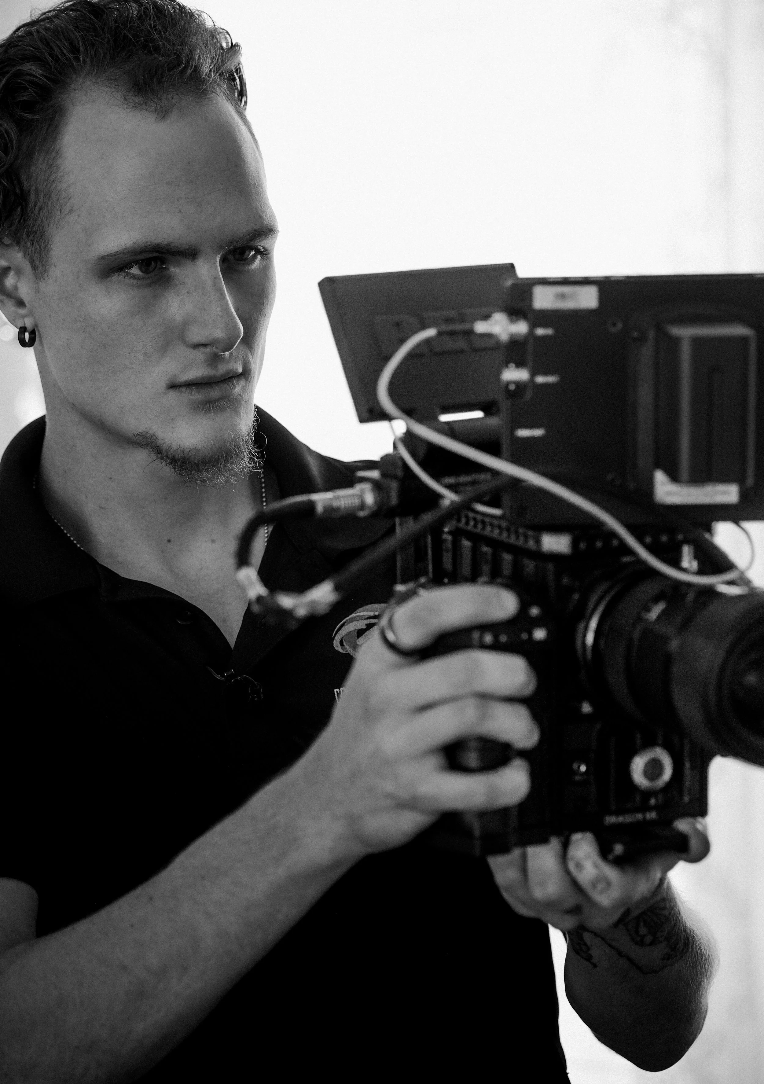 A young man with curly hair and earrings operating a professional camera in a monochrome setting.