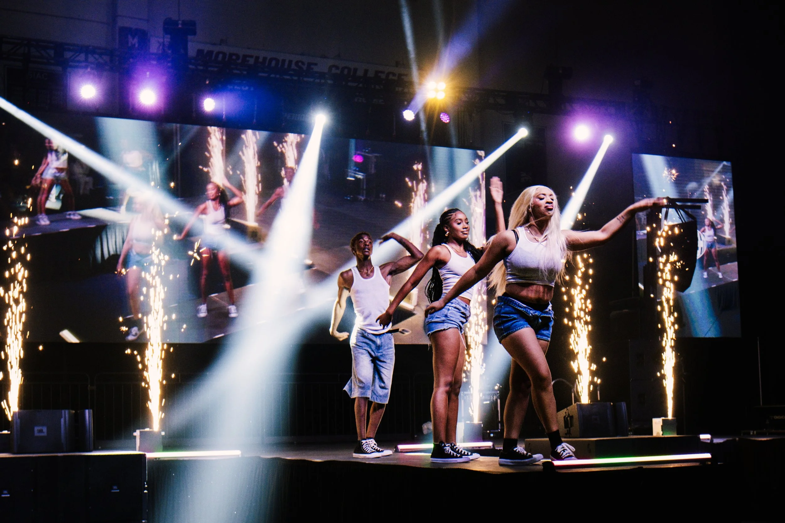 Four young women dancing on stage with pyrotechnics, bright lights, and large screens displaying their movements in the background.