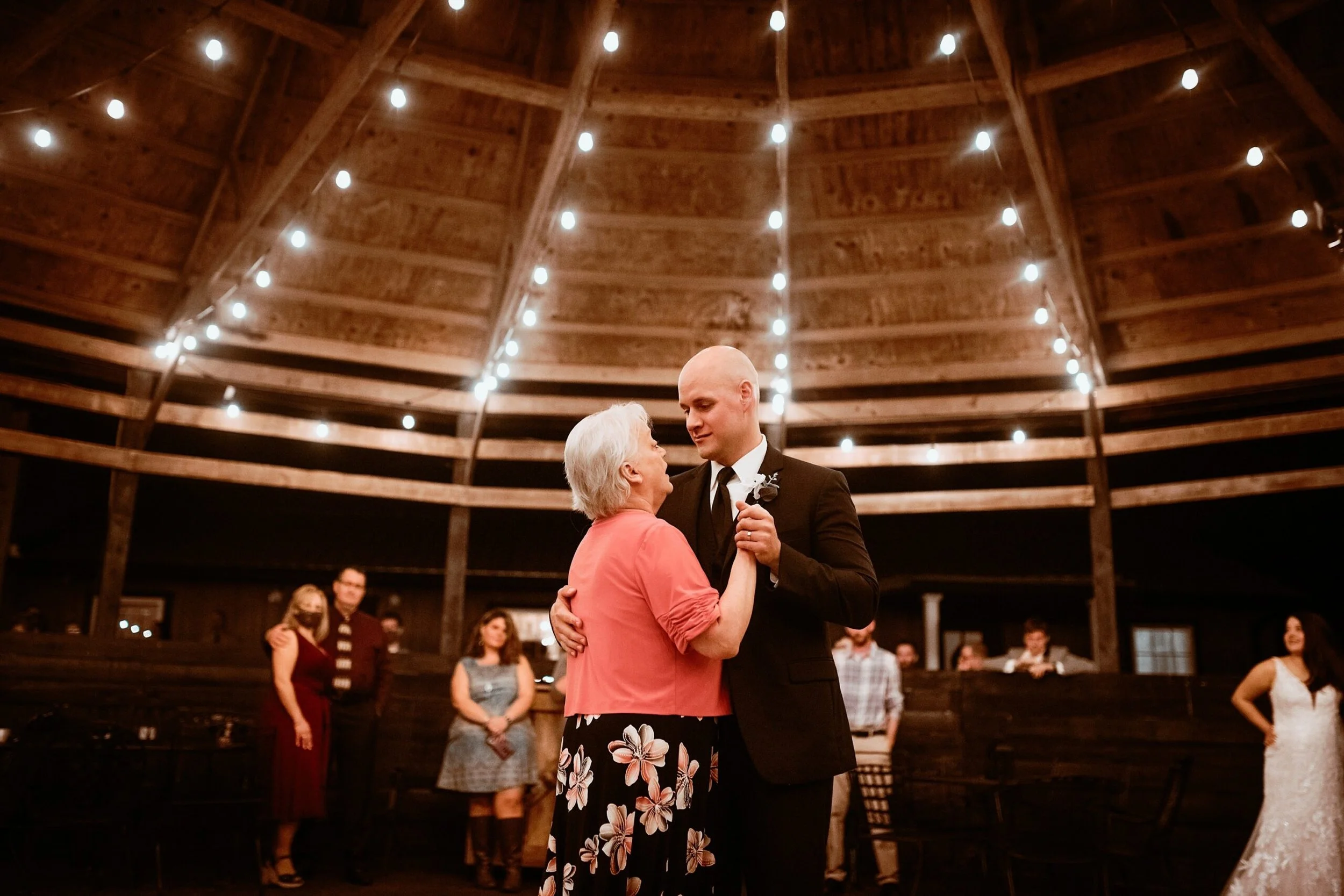 A young man and an older woman dance together at a wedding reception in a rustic barn with string lights overhead. Several guests stand in the background.