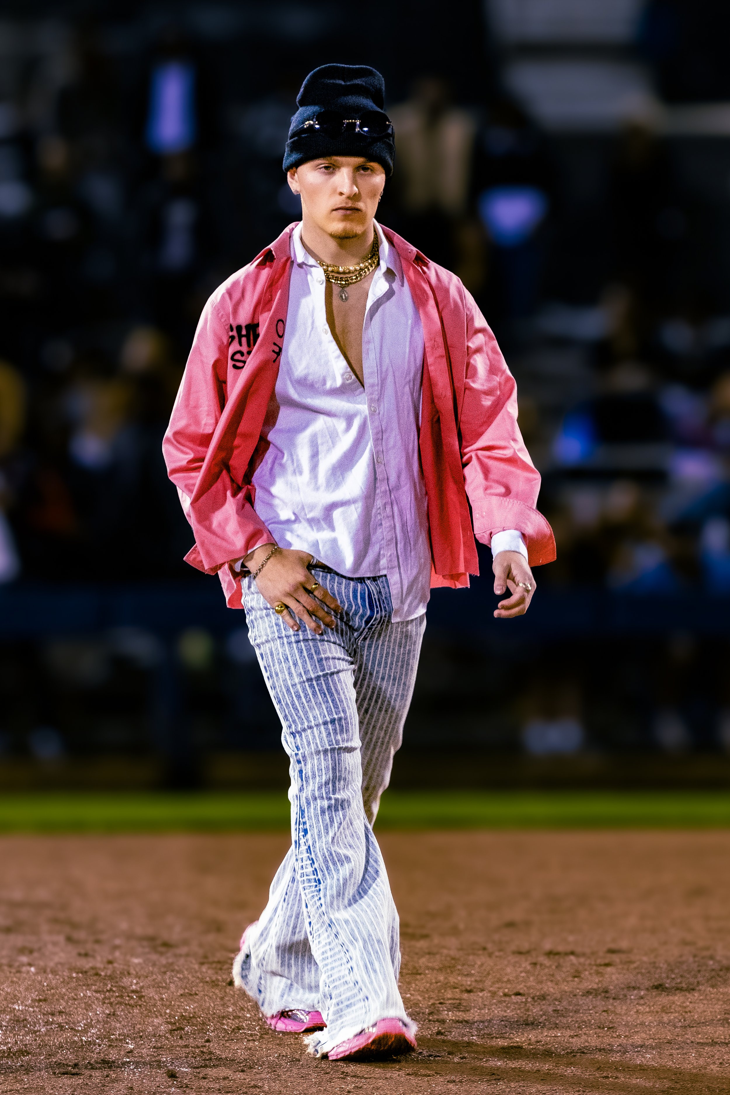 A young man walking on a baseball field at night, wearing a black beanie with sunglasses, a pink jacket, a white shirt, striped pants, pink sneakers, and gold jewelry.