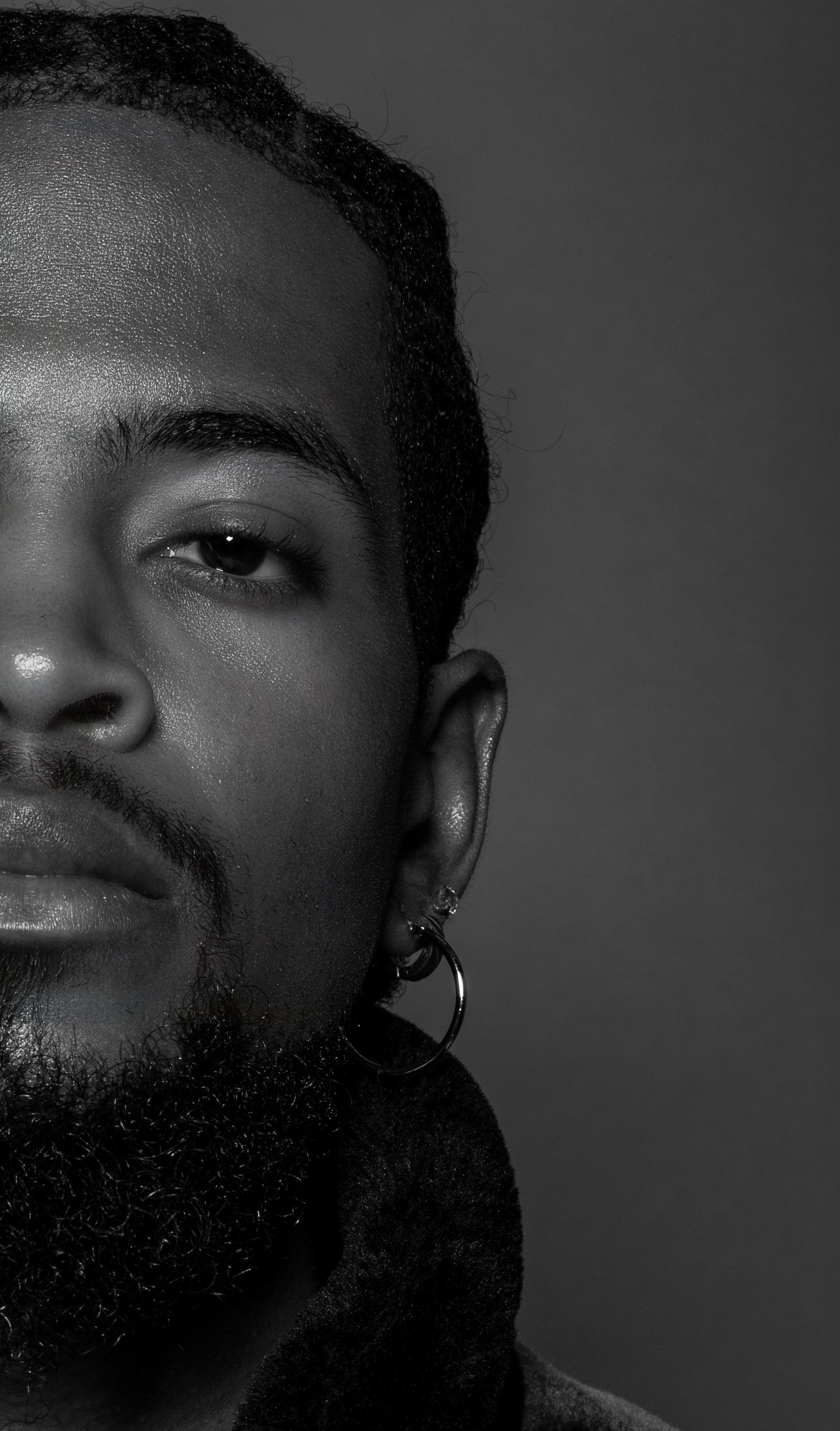 Close-up black and white portrait of a man's face, showing eye, nose, lips, beard, and earrings against a dark background.