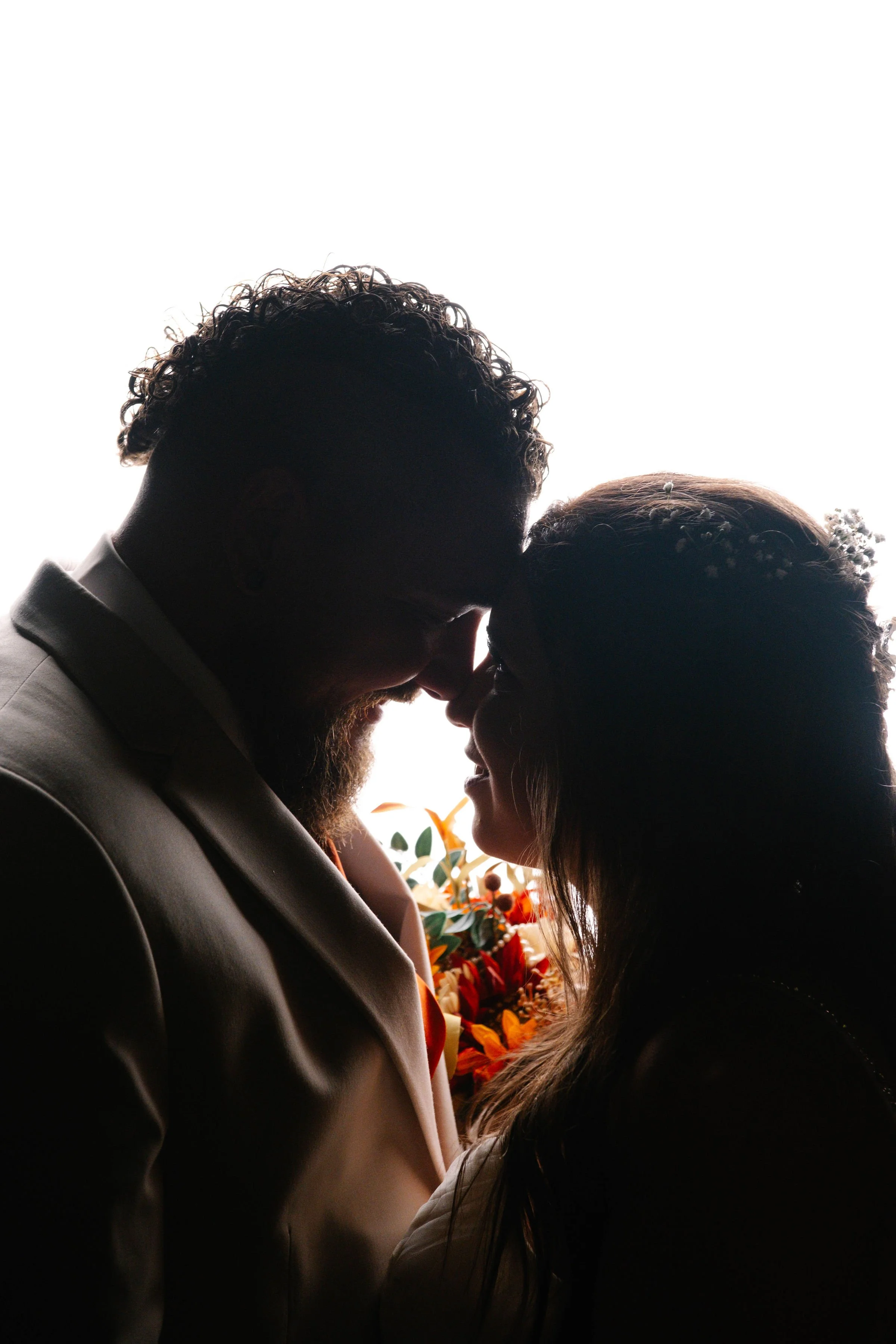A silhouette of a couple touching foreheads with a bright background, floral bouquet visible between them.