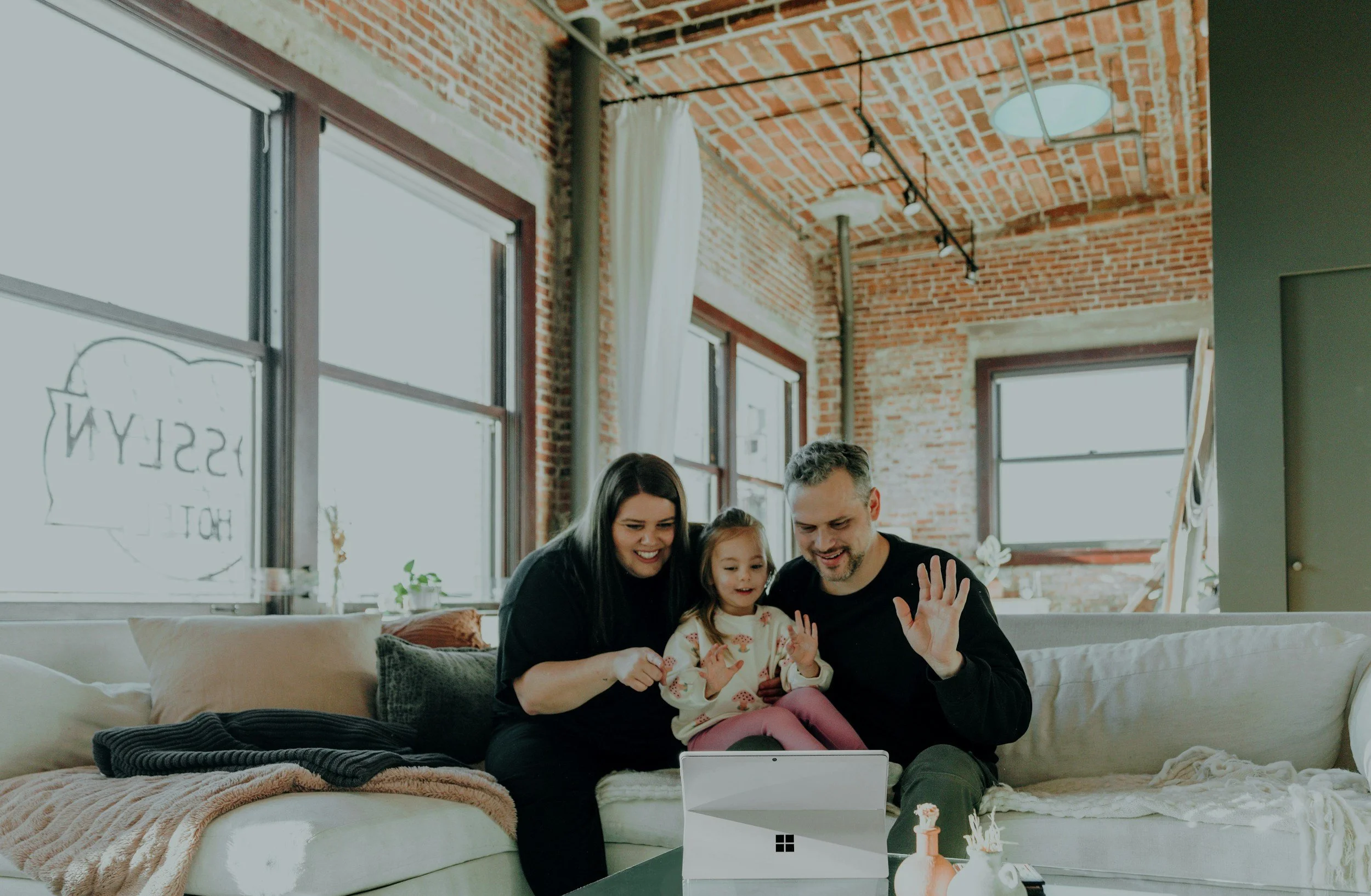 Family of three sitting on a white couch, celebrating and waving at a tablet in a bright, modern living room with large windows, exposed brick walls, and warm decor.