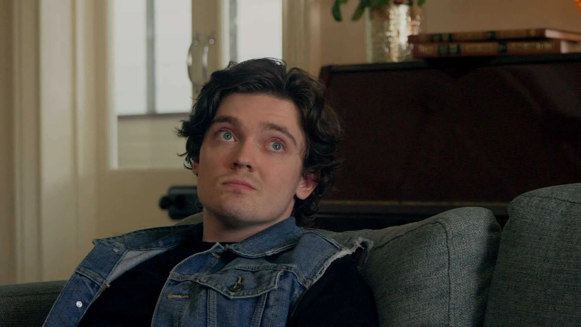 A man with curly black hair and blue eyes looks upward while sitting on a gray sofa, wearing a denim vest over a black shirt in a cozy living room with a window and wooden furniture in the background.