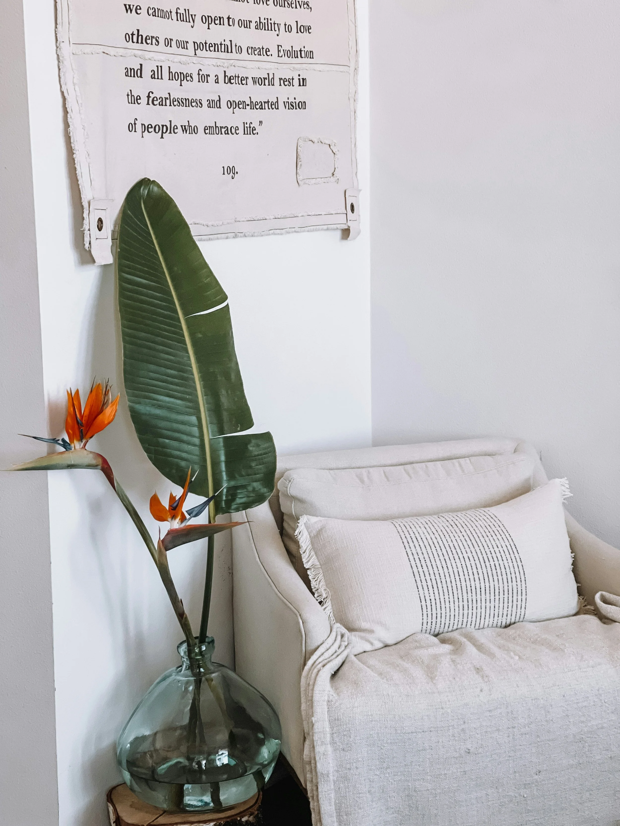 A beige sofa with a decorative pillow, a tall glass vase with large green leaves and orange bird of paradise flowers, and a wall hanging with text.