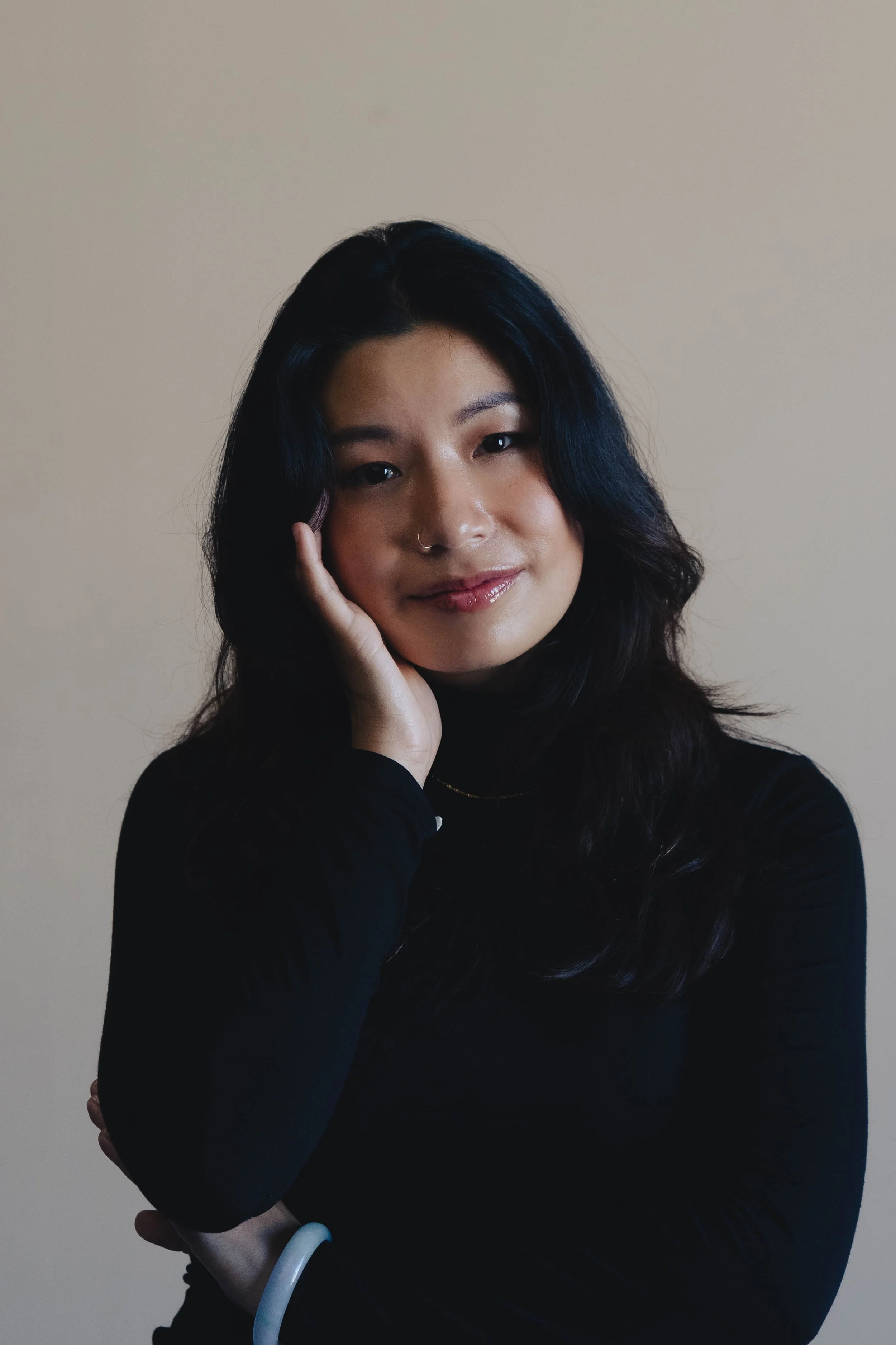 An East Asian person with long wavy black hair, holding their palm to their face, and looking towards the camera with a small smile, wearing a black turtleneck shirt.