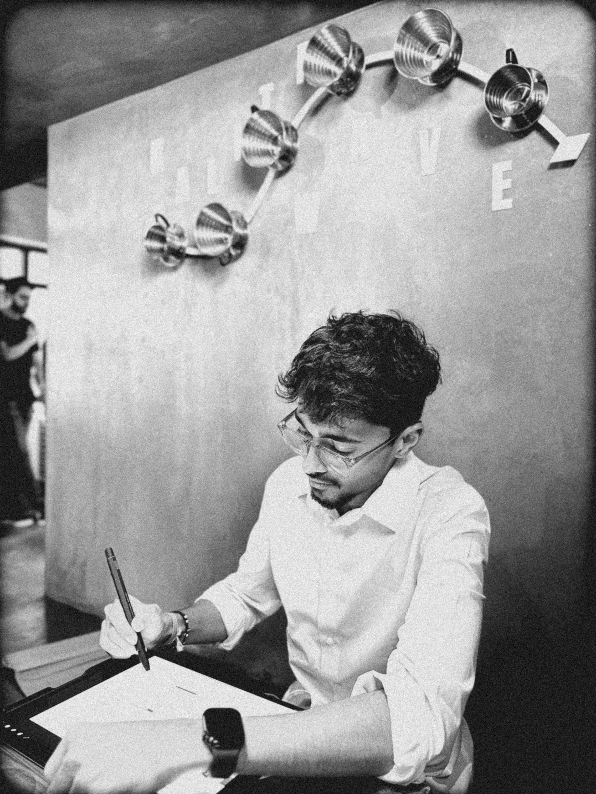 A man with glasses and a white shirt writing or drawing on a large pad at a table, with a wall decorated with metallic balloon art and letters behind him.