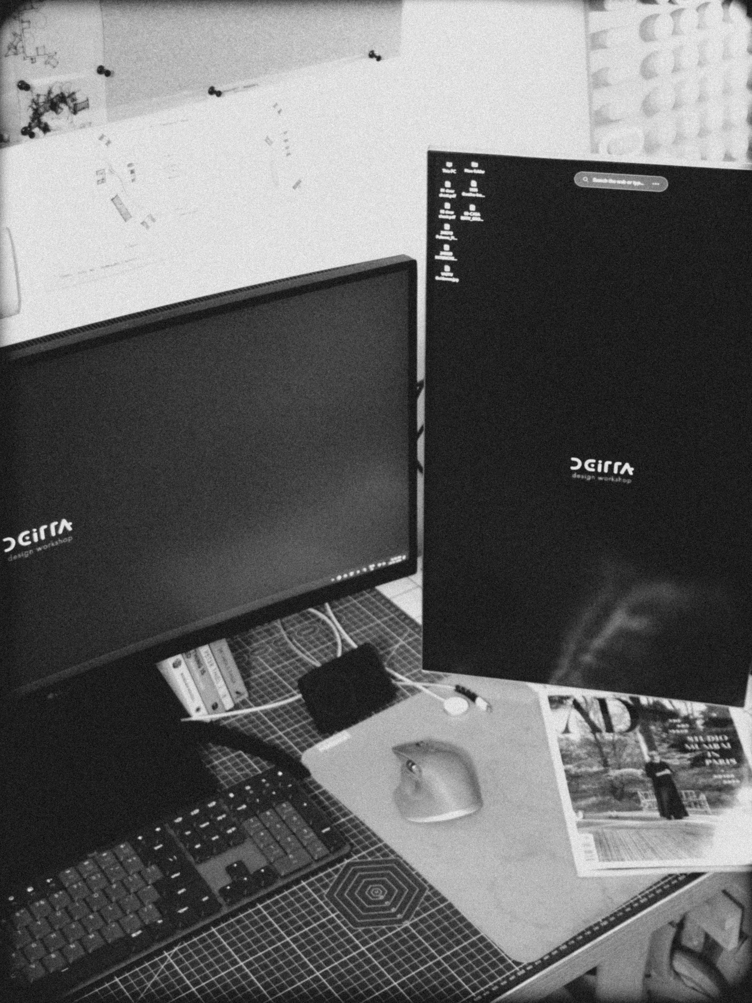 A black and white photo of a workspace with two computer monitors displaying the 'Gira' design workshop logo. The workspace has a mouse, keyboard, a magazine, and some cables on a grid-patterned desk.