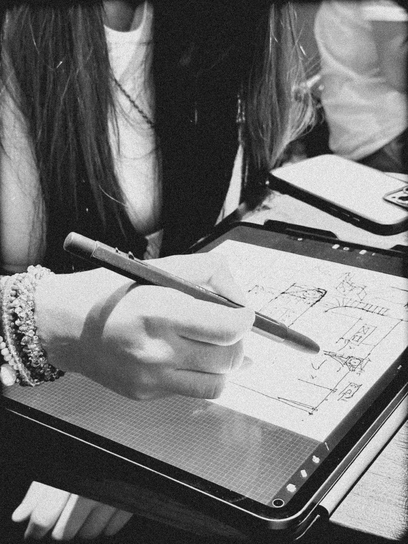 A person with long hair wearing jewelry and a white shirt is drawing on a tablet with a stylus. The drawing appears to be a sketch or blueprint with measurements and annotations.