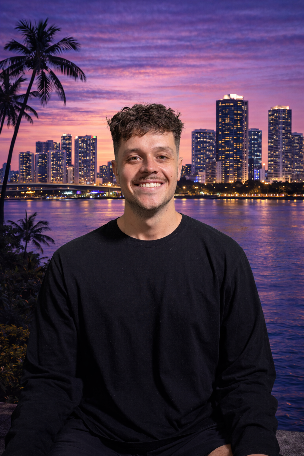 A young man with brown, curly hair smiling in front of a city skyline at dusk, with tall buildings and palm trees reflected in the water.