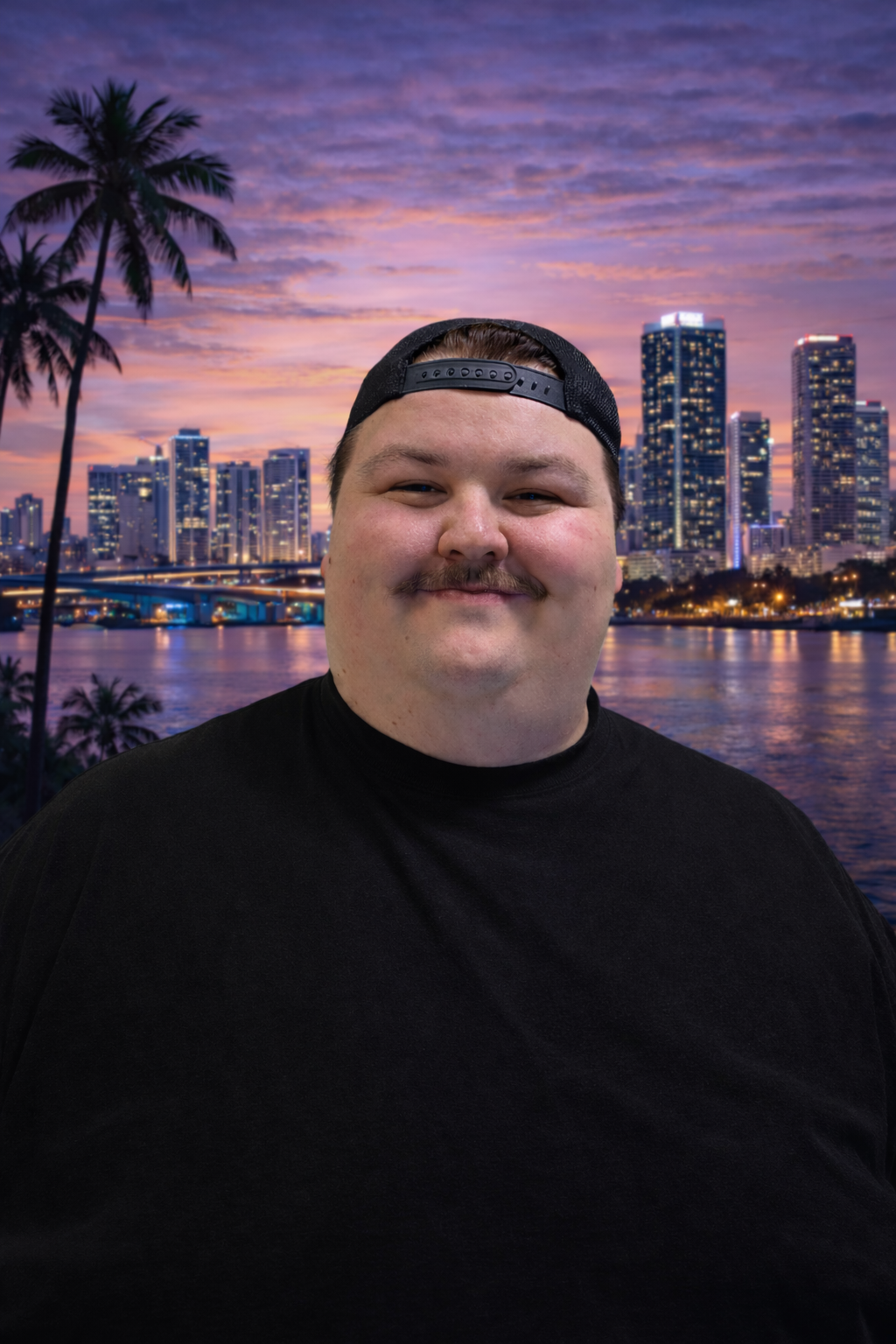 A man with a mustache and wearing a black cap backwards, smiling, with a city skyline and water at sunset in the background.